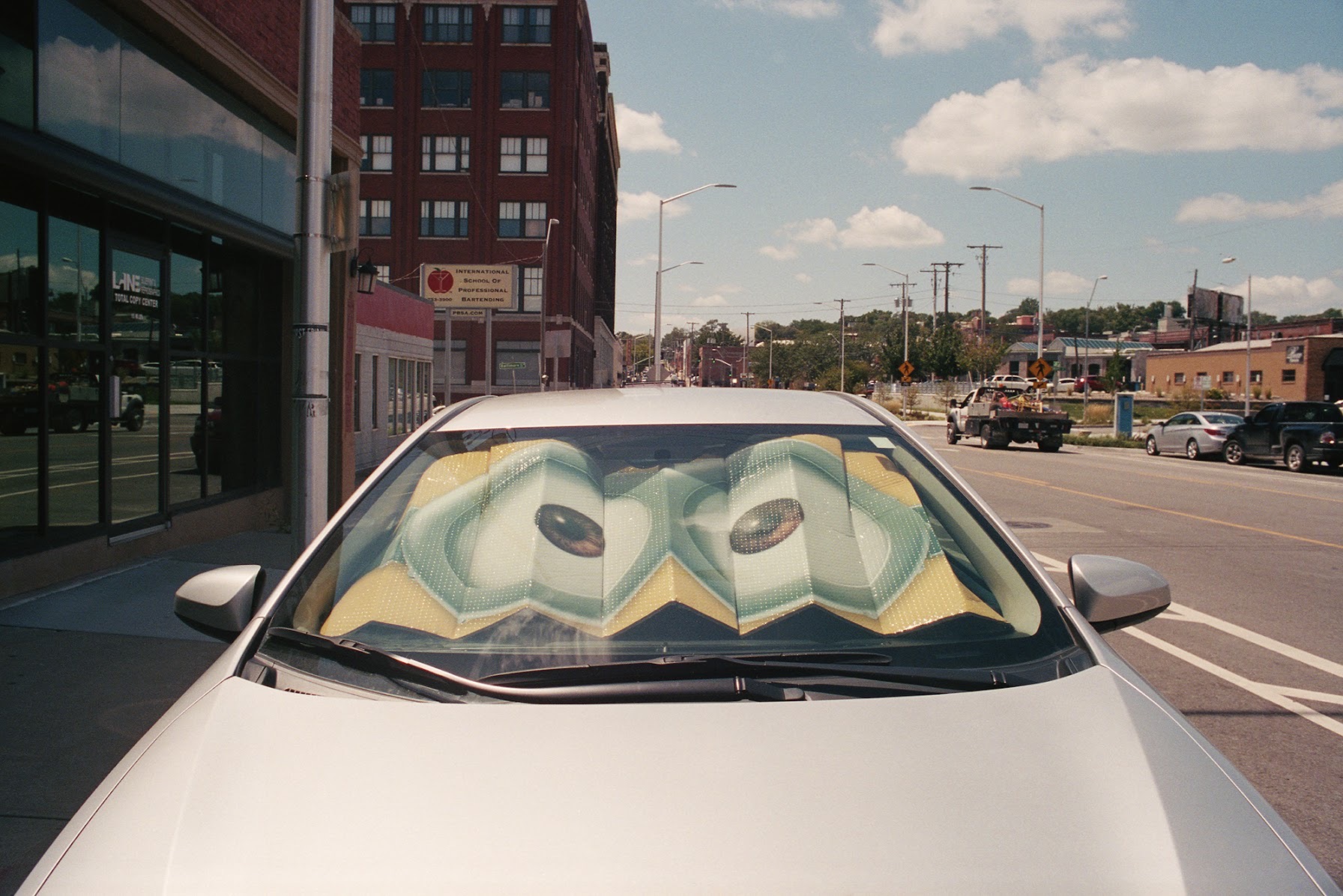 A white car parked on an urban street displays a sun shade featuring a stylized illustration of large eyes with yellow and green coloring, creating an anthropomorphic presence on the vehicle's windshield. Brick buildings, storefronts, and a clear sky line the street behind it.