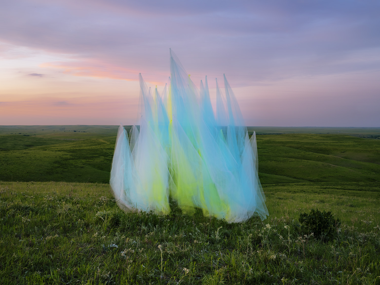 Translucent fabric panels dyed in gradient hues of white, yellow, and blue stand vertically in an open grassland under a soft sunset sky, creating an ethereal sculptural form against the rolling prairie landscape.