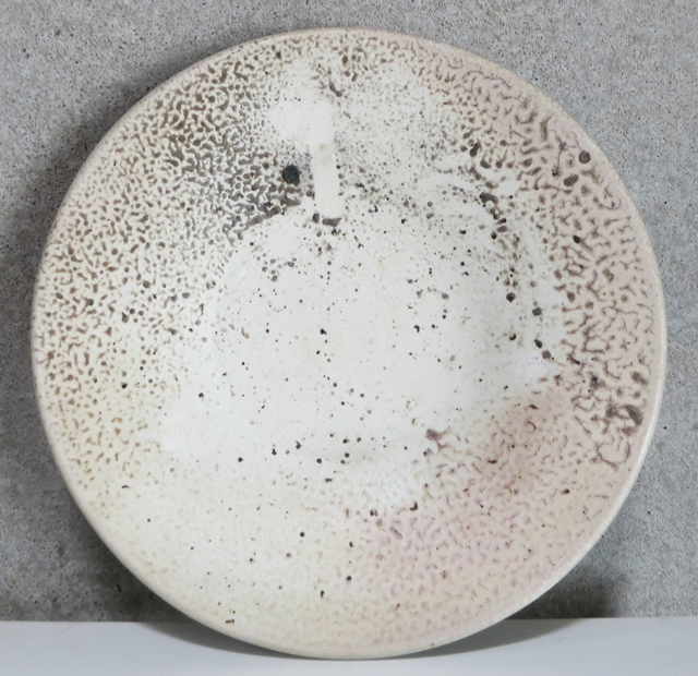 A circular porcelain platter with a speckled cream and taupe surface featuring organic, textured patterning around its rim and a smooth, minimally marked center. The wood-fired surface displays natural variations in color and subtle ash deposits characteristic of high-temperature firing.