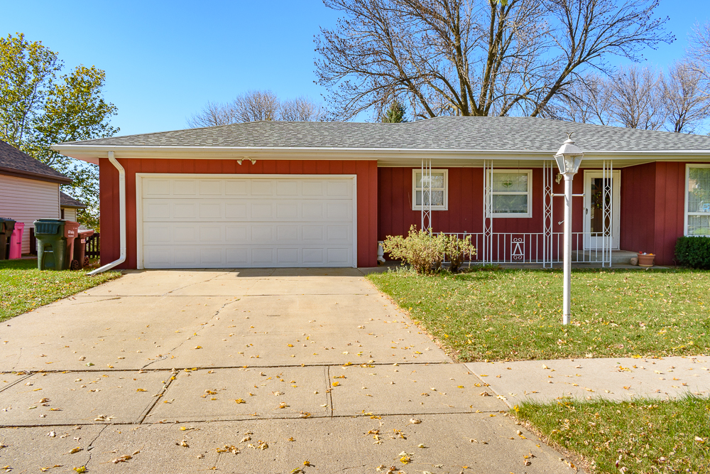 A red-brick residential house with a white garage door and covered porch, photographed from the street on a clear day. Bare and partially leafed trees stand in the background beneath a blue sky, while fallen leaves scatter across the concrete driveway and lawn in the foreground.