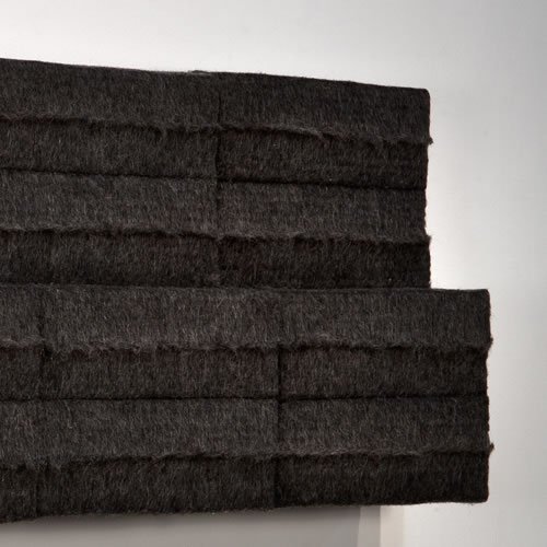 Stacked layers of dark charcoal felt arranged in an angular, stepped composition against a white wall, with visible horizontal creases and a tactile, textured surface that creates subtle shadows and depth.