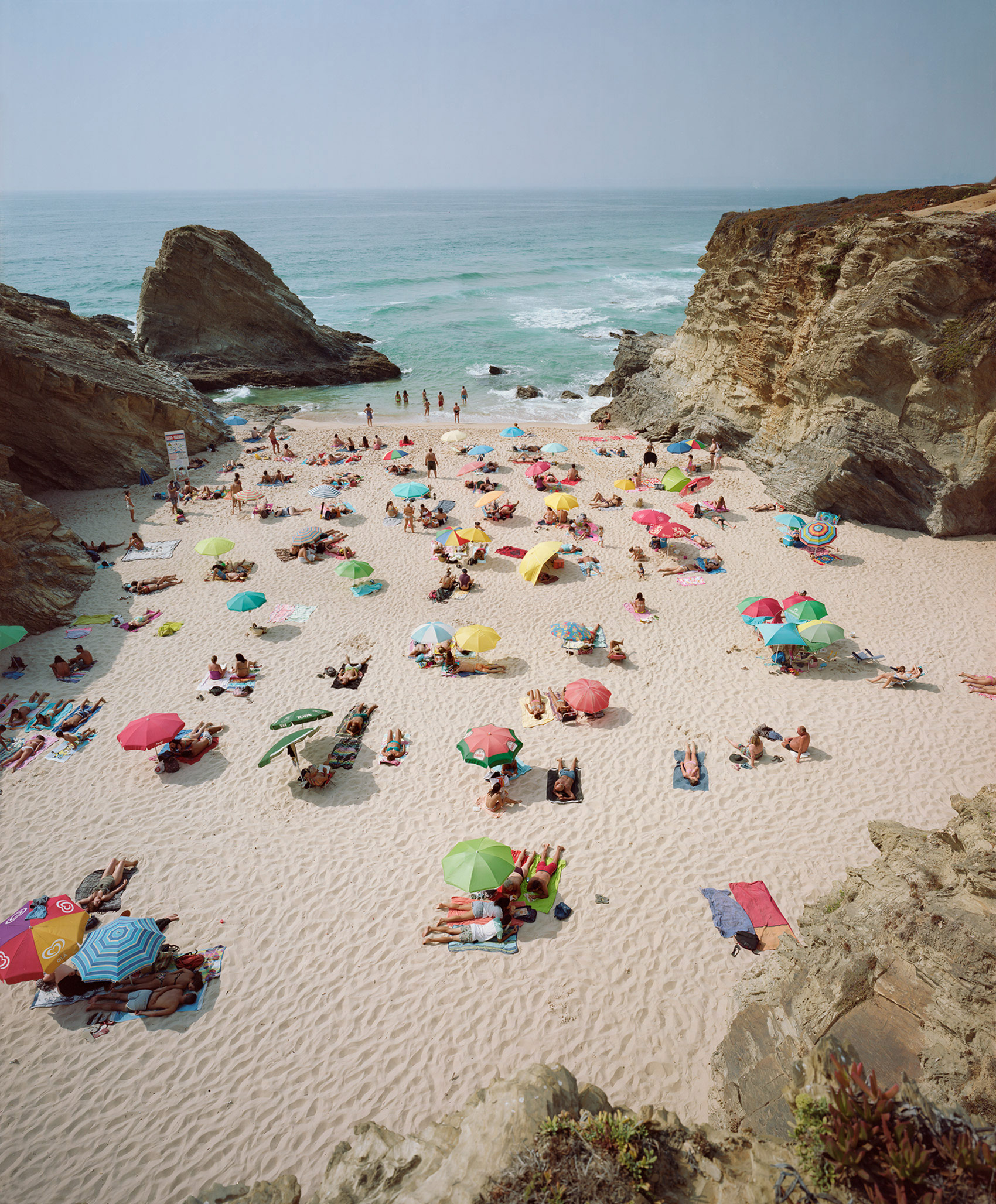 Aerial view of a crowded cove beach framed by dramatic limestone cliffs, with sunbathers scattered across pale sand beneath a vibrant array of multicolored umbrellas in pink, green, yellow, and blue; turquoise waters and rocky outcroppings visible beyond the shoreline.
