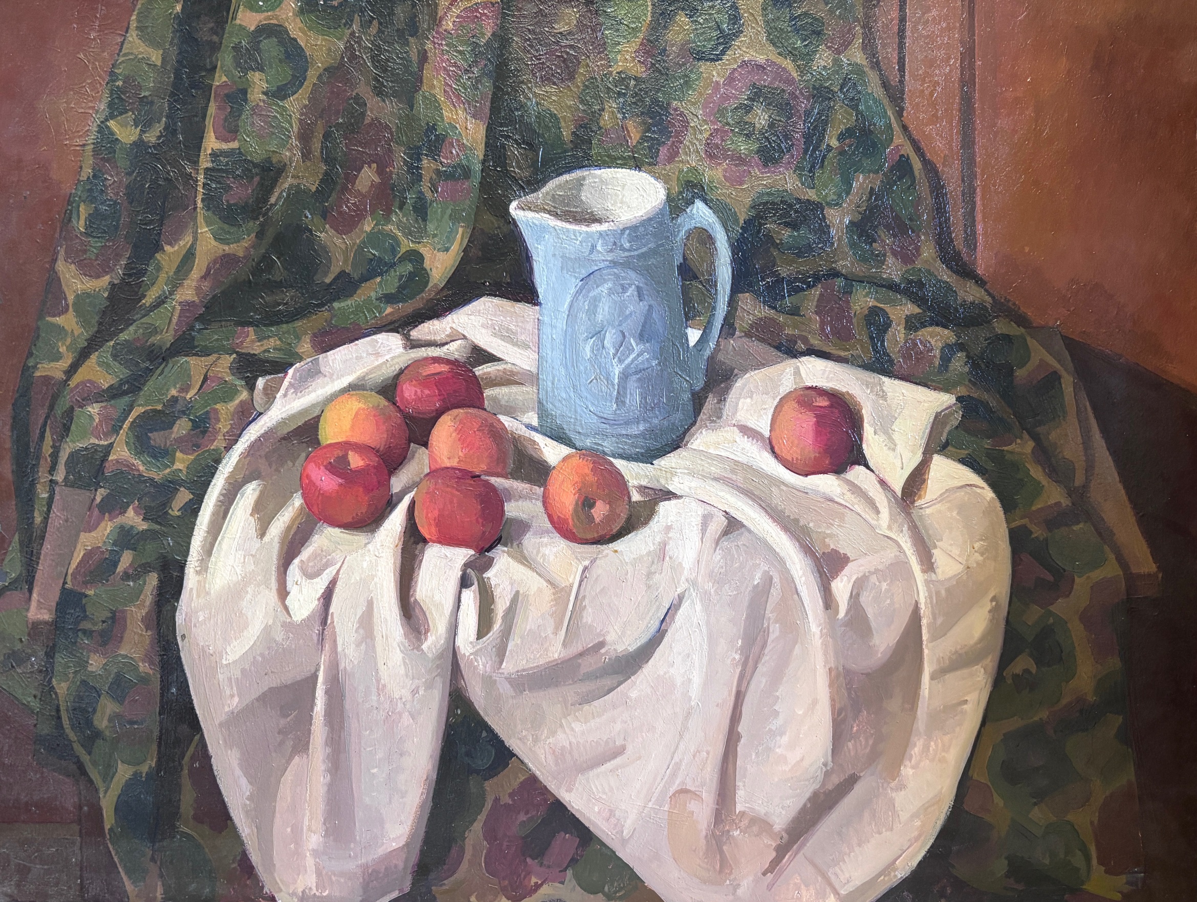 A still life composition featuring red apples arranged on white draped fabric, with a blue-grey pitcher positioned behind them. The arrangement sits against a richly patterned backdrop of dark fabric with multicolored floral motifs, set within a warm reddish-brown interior space.