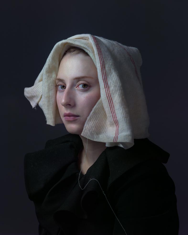 Portrait of a young woman against a dark background, wearing a black garment and a cream-colored linen head covering with a red stripe. Her gaze is directed slightly away from the camera, with a composed, contemplative expression.