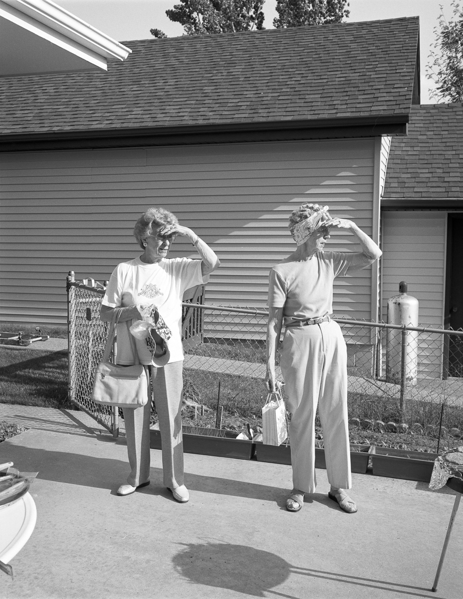 Black and white photograph showing two women standing in a residential backyard, both adjusting their sunglasses or visors with their hands raised to their heads. They stand on a concrete patio beside a chain-link fence, with a single-story house and shingled roof visible behind them. Various household items and garden tools are scattered around the yard.