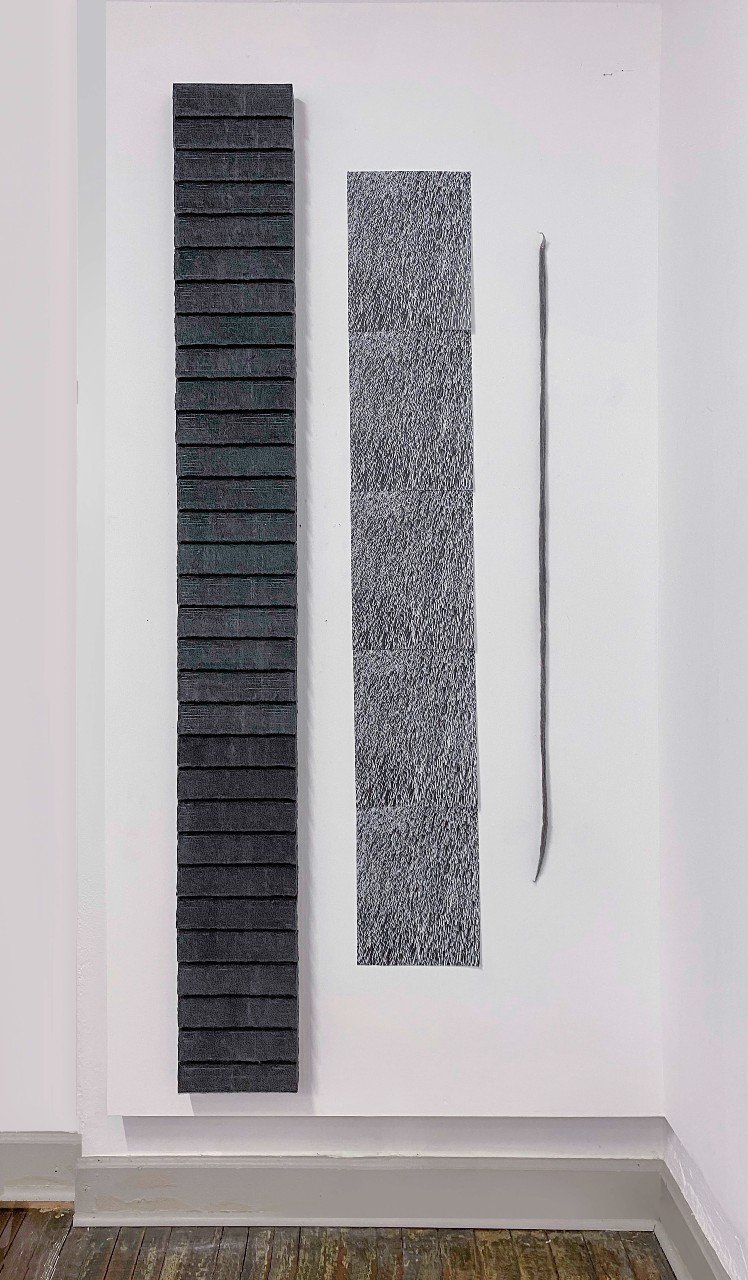 Three vertical textile artworks mounted on a white gallery wall: a tall narrow strip of dark gray wool felt with horizontal divisions, a textured gray rectangular panel with dense linear markings, and a thin curved metallic line element, displayed against a wooden floor.