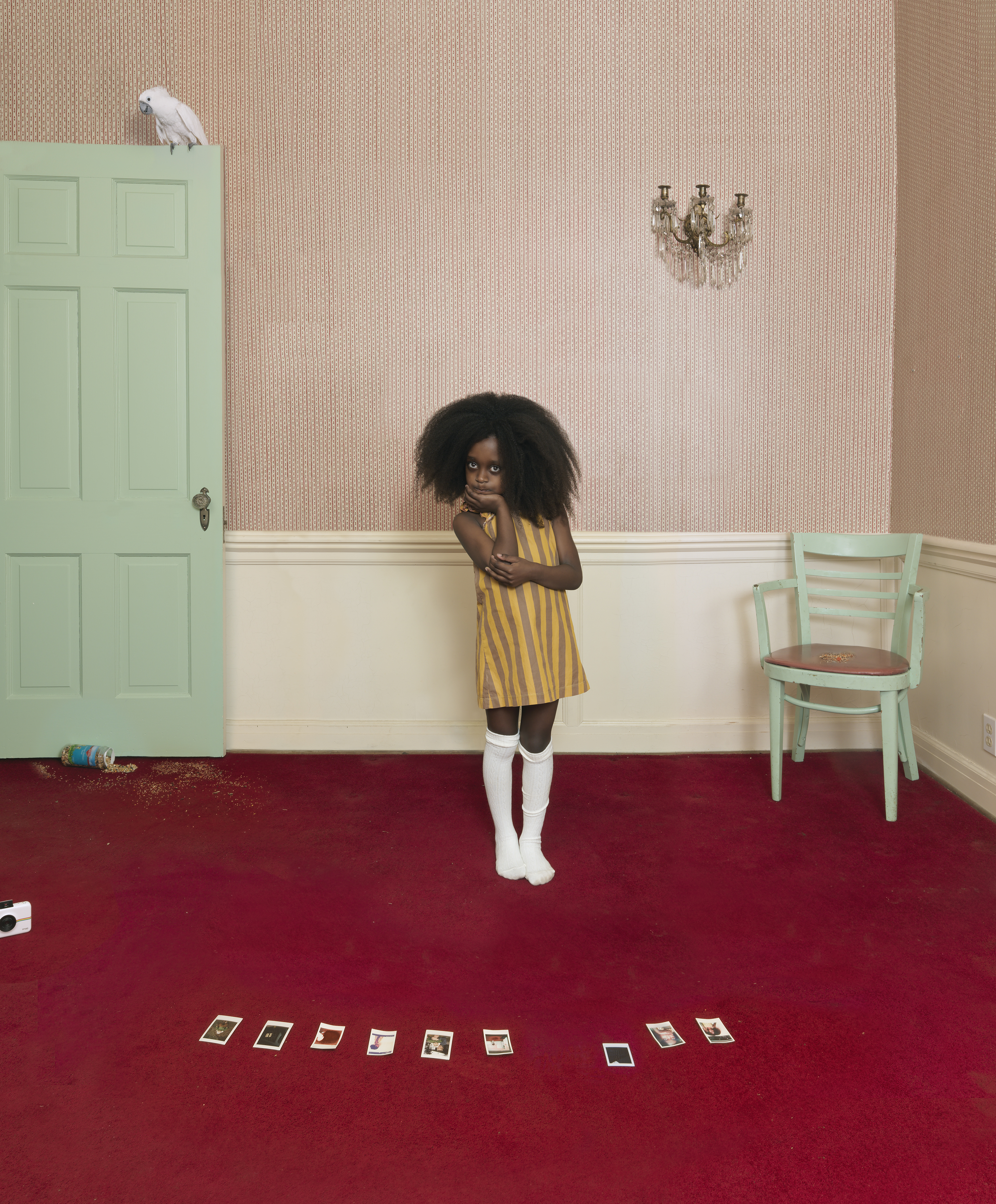 A young child with voluminous dark hair stands in the center of a formally decorated interior room, wearing a mustard-yellow striped dress and white socks. The room features soft pink textured wallpaper above a cream-colored wainscot, a mint green door and chair, ornate wall sconce, and deep red carpet scattered with small photographs or cards arranged in linear patterns across the floor.