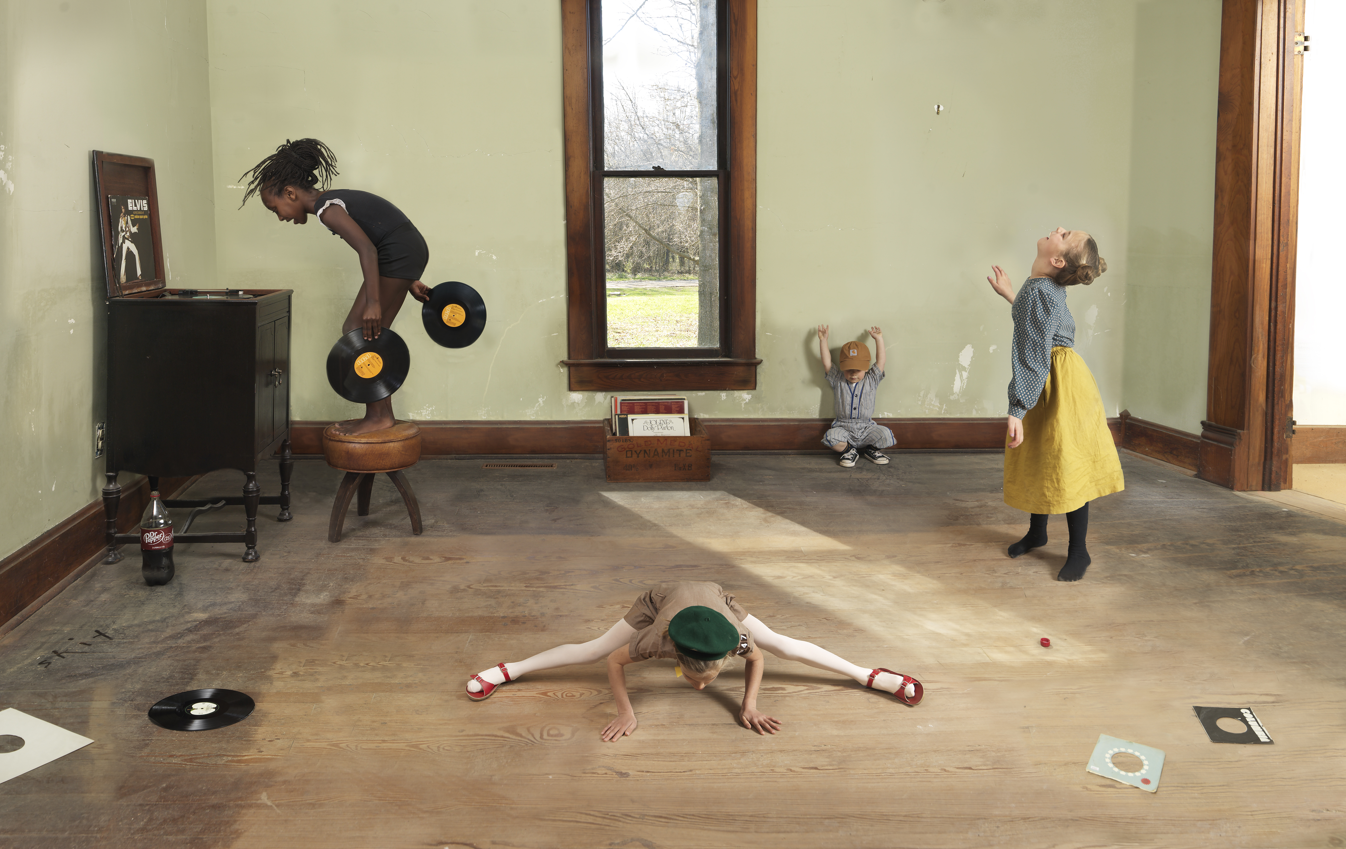 Four figures arranged throughout a pale green-walled room engage with vinyl records as sculptural and performative objects. A person in a black shirt balances on a wooden stool while holding records, another lies horizontally on the floor in a split stretch wearing a green beret, a small child sits in the center, and a figure in a yellow skirt stands to the right with arms raised. Vinyl records and related ephemera are scattered across the wooden floor, while a window frames bare trees outside.