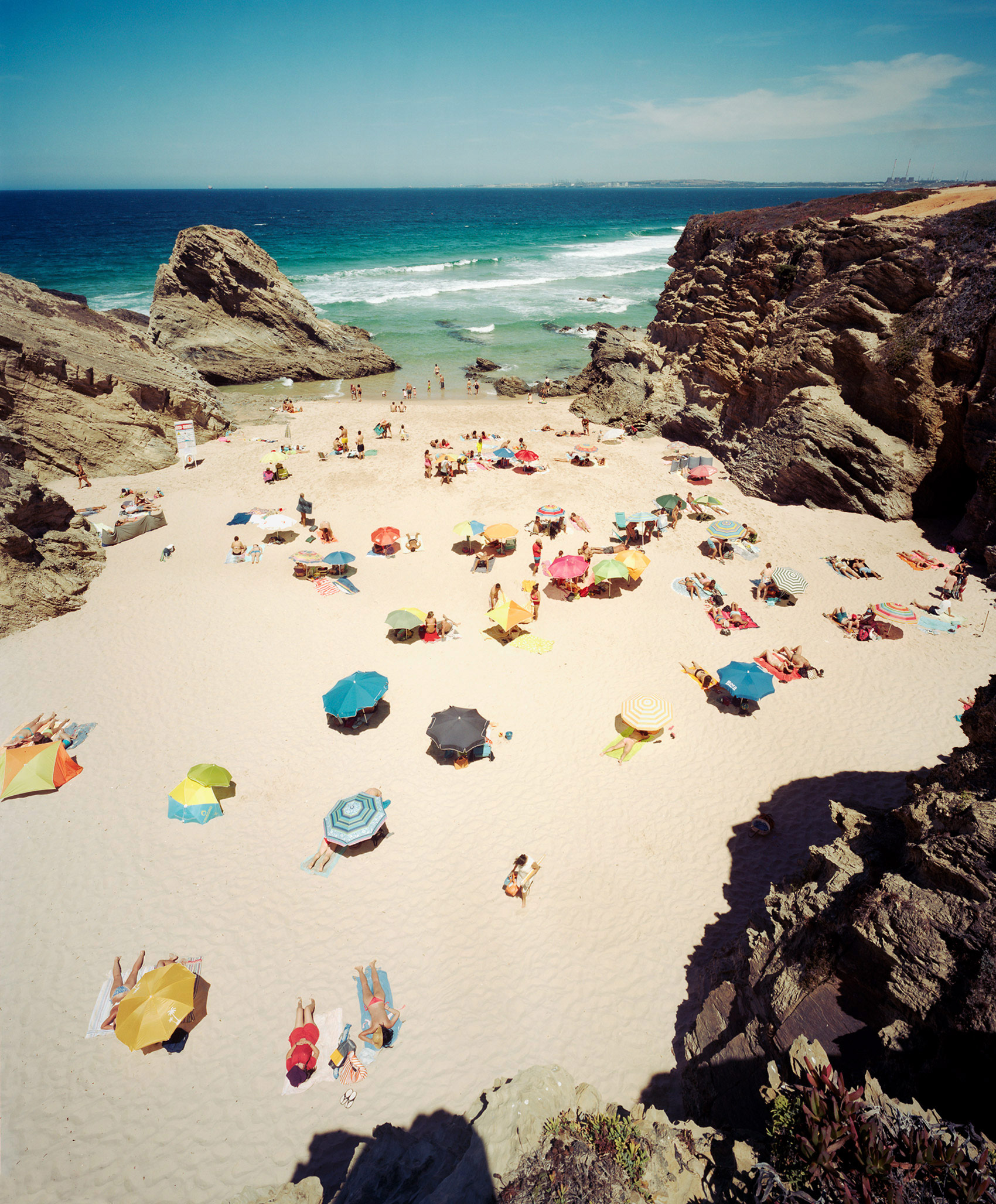 Aerial view of a crowded beach cove enclosed by dramatic rocky cliffs, with numerous colorful umbrellas scattered across pale sand and turquoise waters dotted with swimmers and small waves beyond.