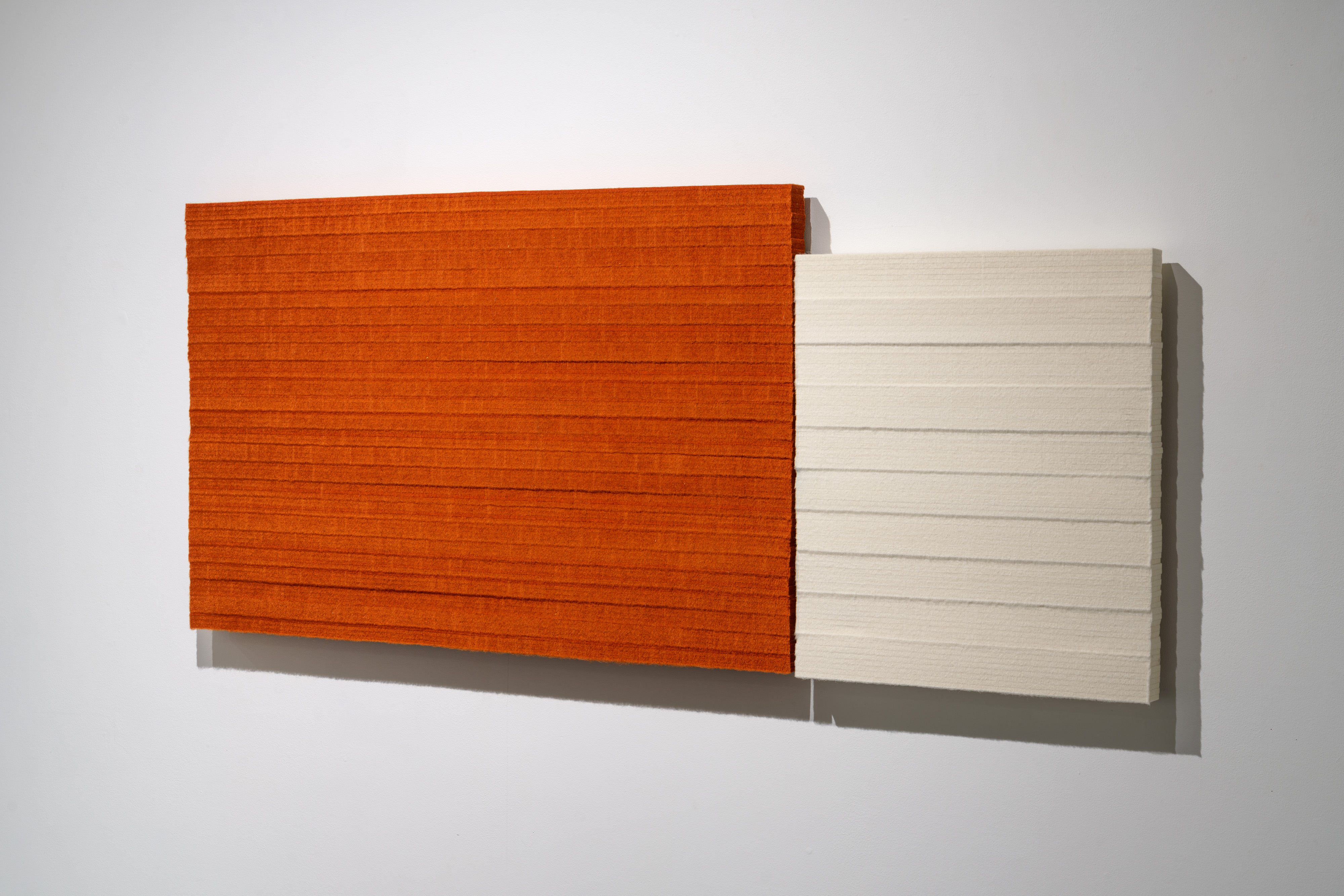 Two rectangular panels mounted horizontally on a white wall—one vibrant burnt orange on the left, one cream-colored on the right—both displaying subtle horizontal striations across their surfaces and casting soft shadows beneath.