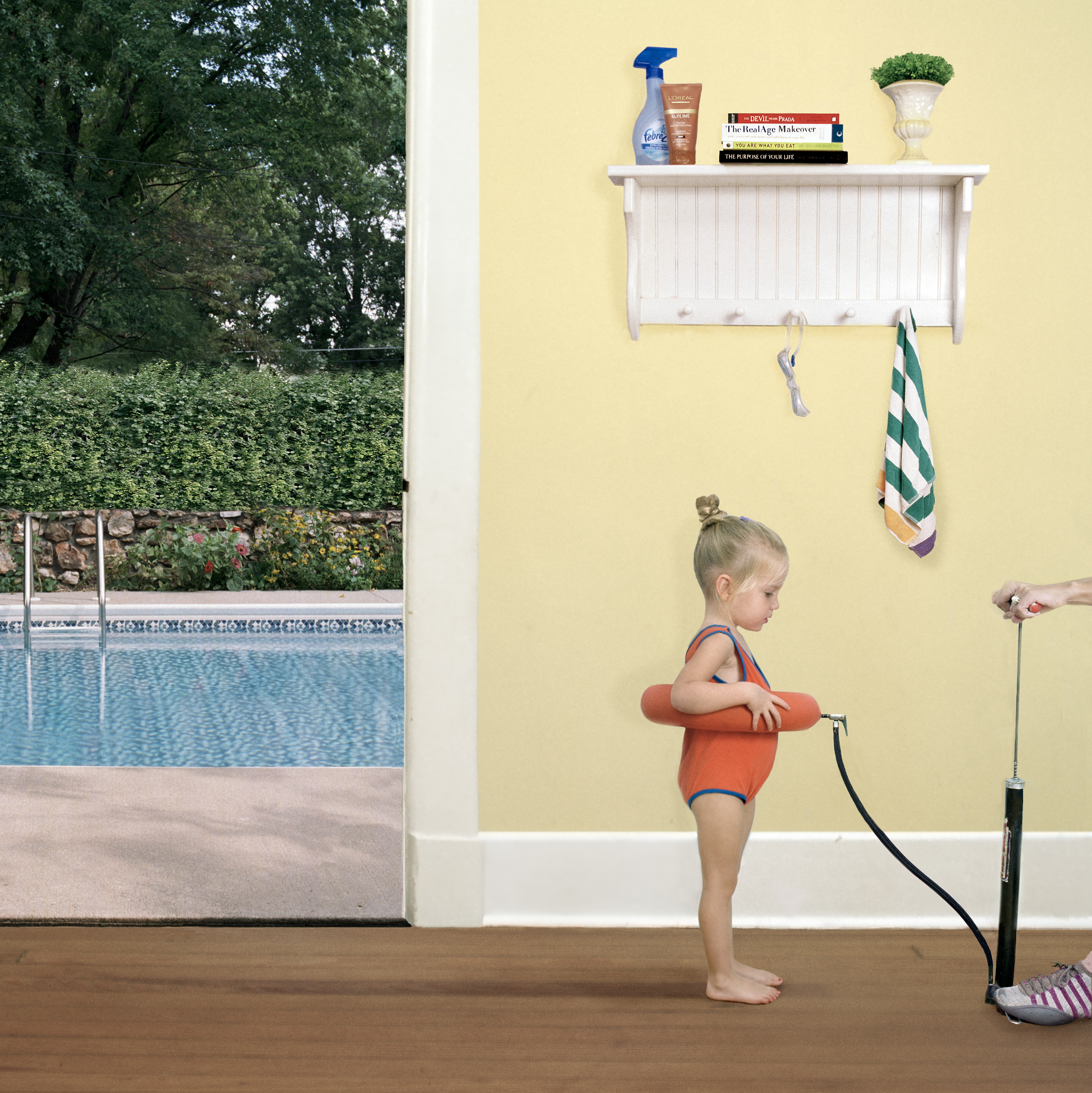 A young child wearing an orange swim floatie stands on a wooden floor facing a pale yellow wall, gazing toward an open glass door that frames a backyard swimming pool surrounded by green hedges. A white shelf mounted on the wall above holds books and household items, with towels and a hand pump hanging below, while pool maintenance equipment sits on the floor beside the child.