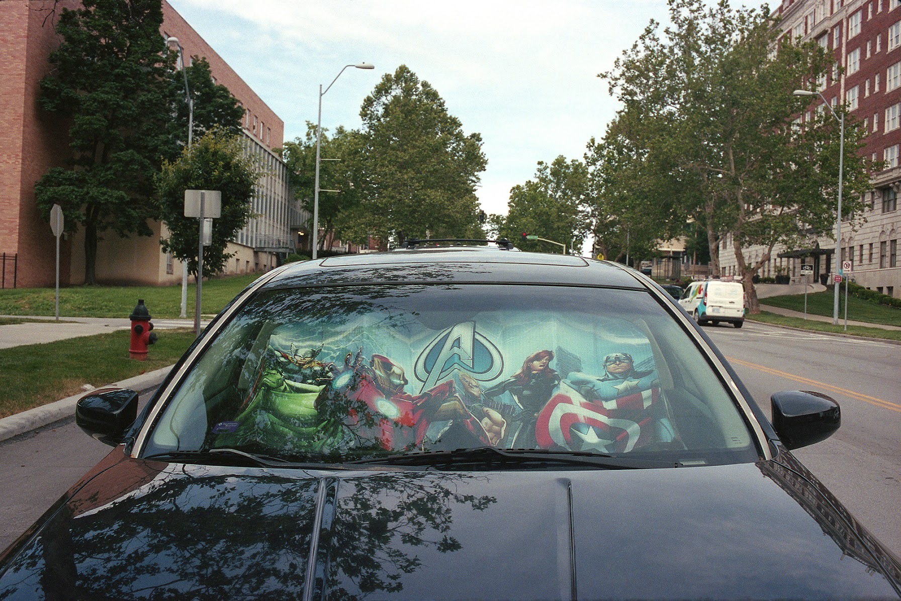 A car's front windshield displays a large-scale printed image of Marvel's Avengers characters in dynamic poses, rendered in vibrant blues, greens, and reds. The artwork is photographed head-on as the vehicle sits parked on an urban street lined with brick buildings and trees.