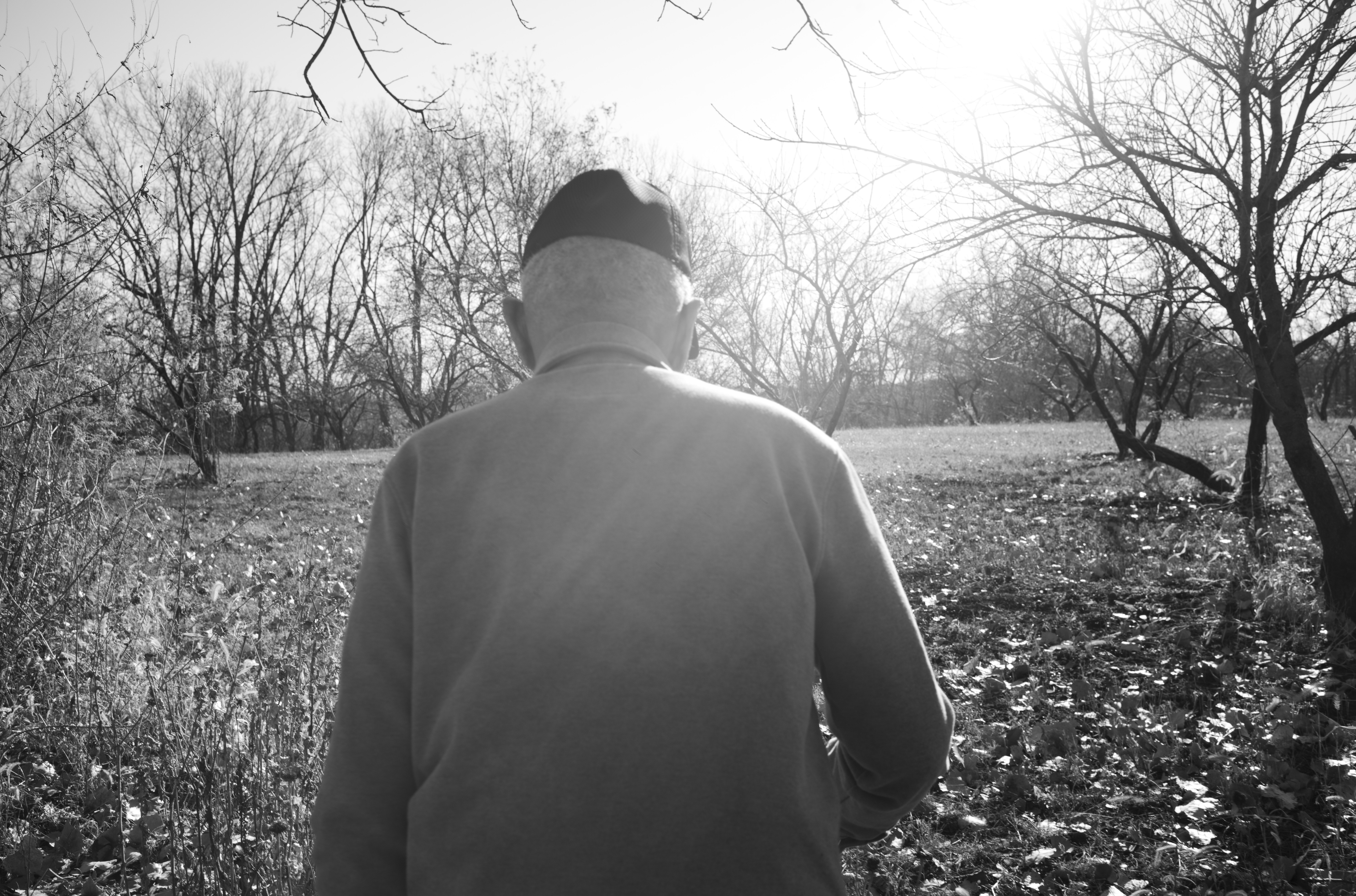 Black and white photograph showing a solitary figure viewed from behind, wearing a light-colored jacket and dark cap, standing in a sparse winter landscape bordered by bare trees. The composition creates a sense of isolation and contemplation, with the figure gazing toward an open, desolate field.