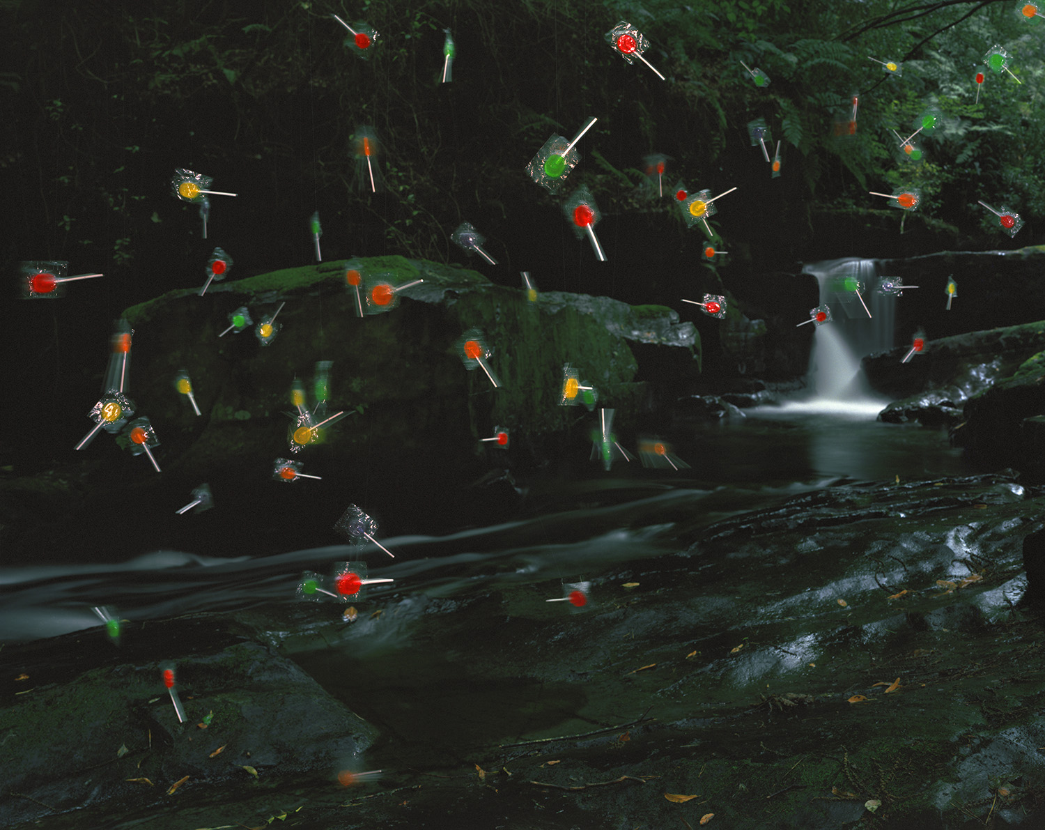 A serene forest stream with moss-covered rocks and a small waterfall is populated by dozens of colorful lollipops with red, orange, yellow, and green spherical heads and white sticks, floating and positioned throughout the scene as if suspended in the air and landscape.