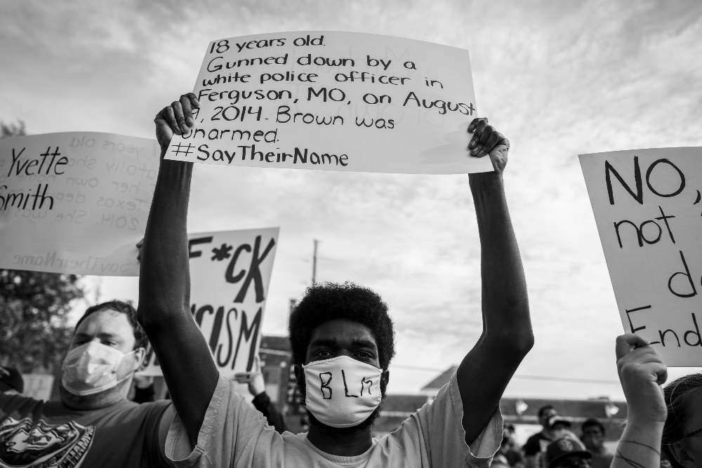 Black and white photograph of a protest scene showing a person wearing a white mask with "BLM" written on it, holding a handwritten sign above their head documenting the death of Michael Brown in Ferguson, Missouri. Multiple other protest signs are visible in the composition, with crowds gathered in the background under an overcast sky.