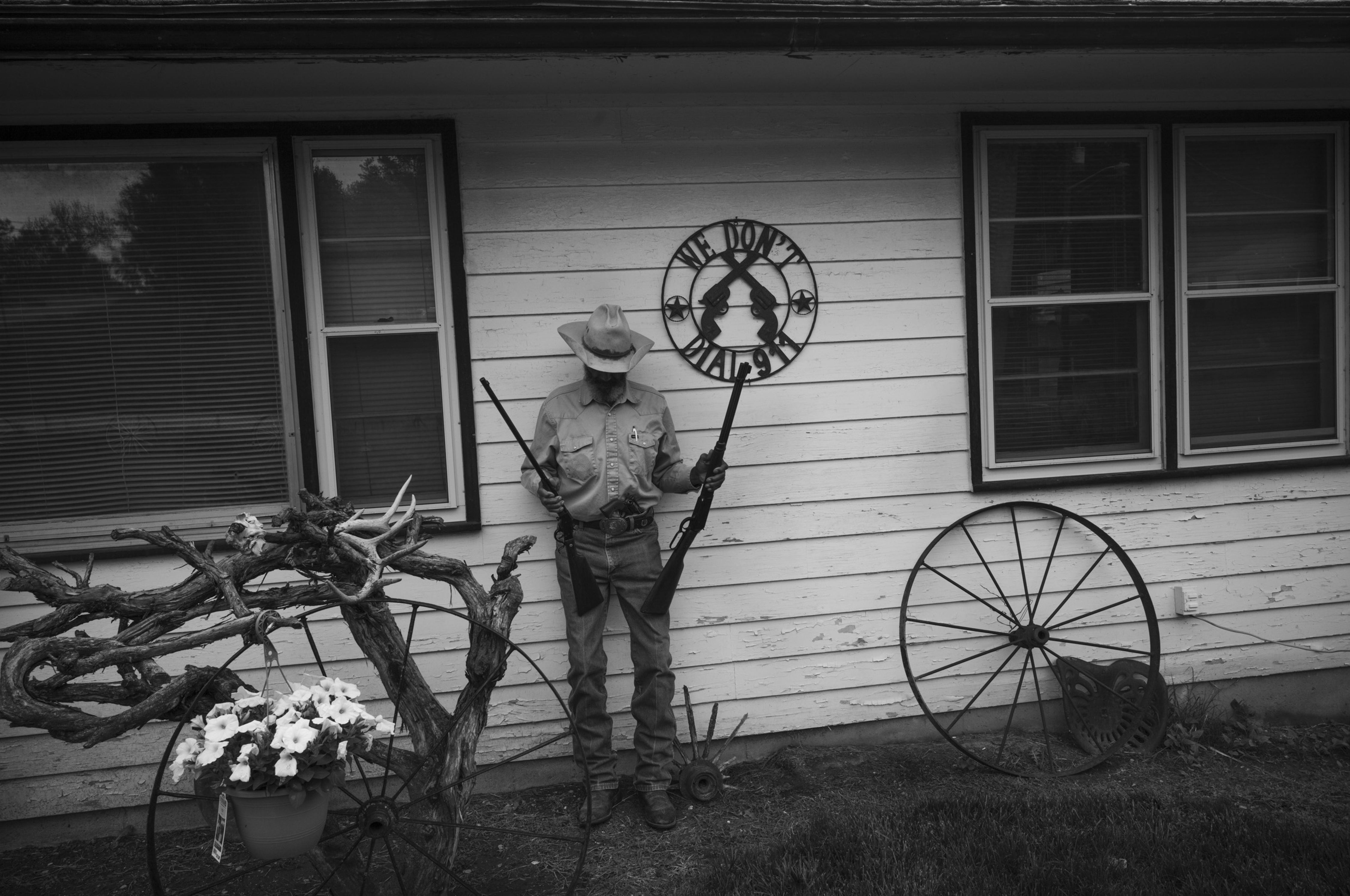 Black and white photograph of a park ranger in uniform and wide-brimmed hat standing against a wooden building wall, holding an ornate metal sign above his head. Rustic objects surround him including wagon wheels, antlers, and a potted plant with white flowers, arranged as if in a folk art or Western-themed display.
