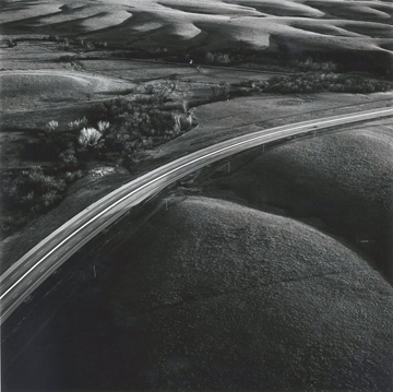 Aerial black and white photograph of a curved highway cutting through rolling prairie landscape, with agricultural fields visible in the distance and a cluster of trees and buildings nestled in a valley alongside the road.