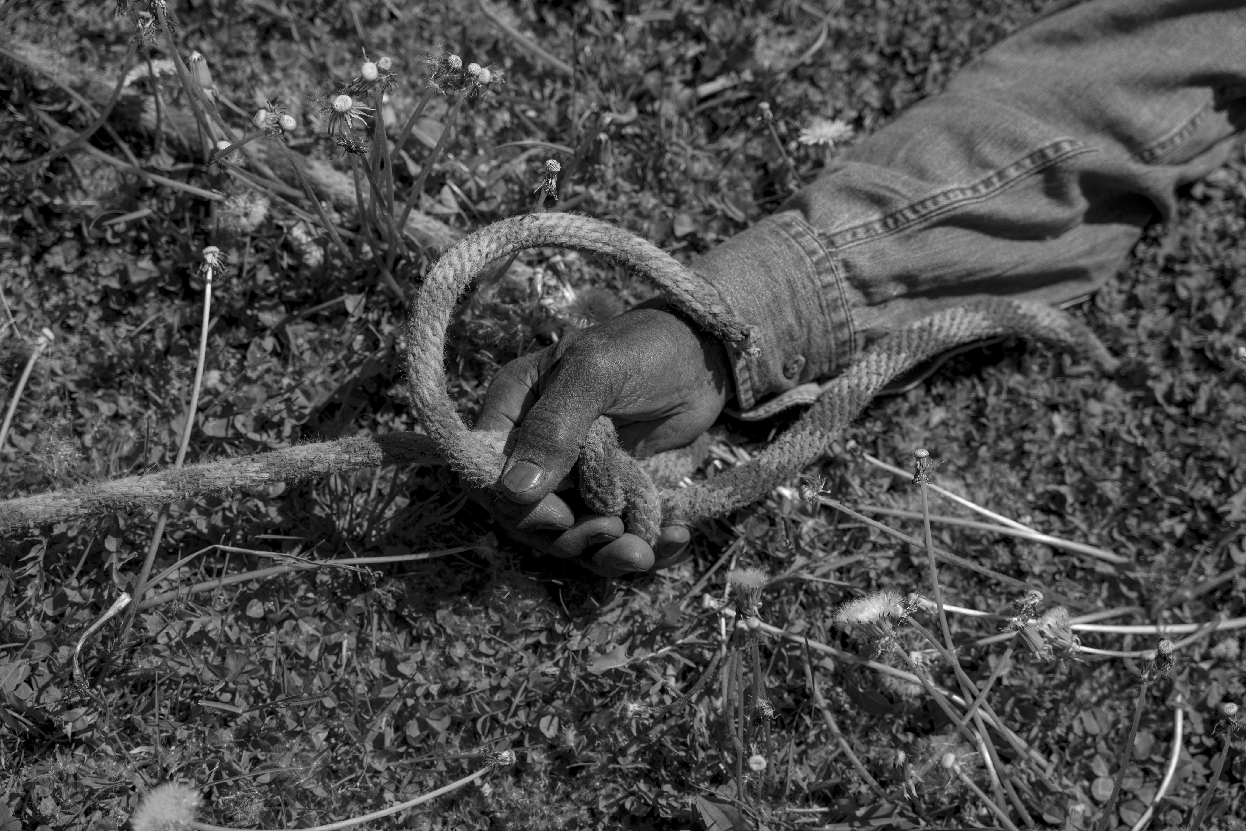 Black and white photograph of a hand wearing a textured glove, grasping a coiled rope against a ground of wildflowers and dried vegetation. The composition captures an intimate moment between human gesture and natural landscape, shot from above with shallow depth of field.