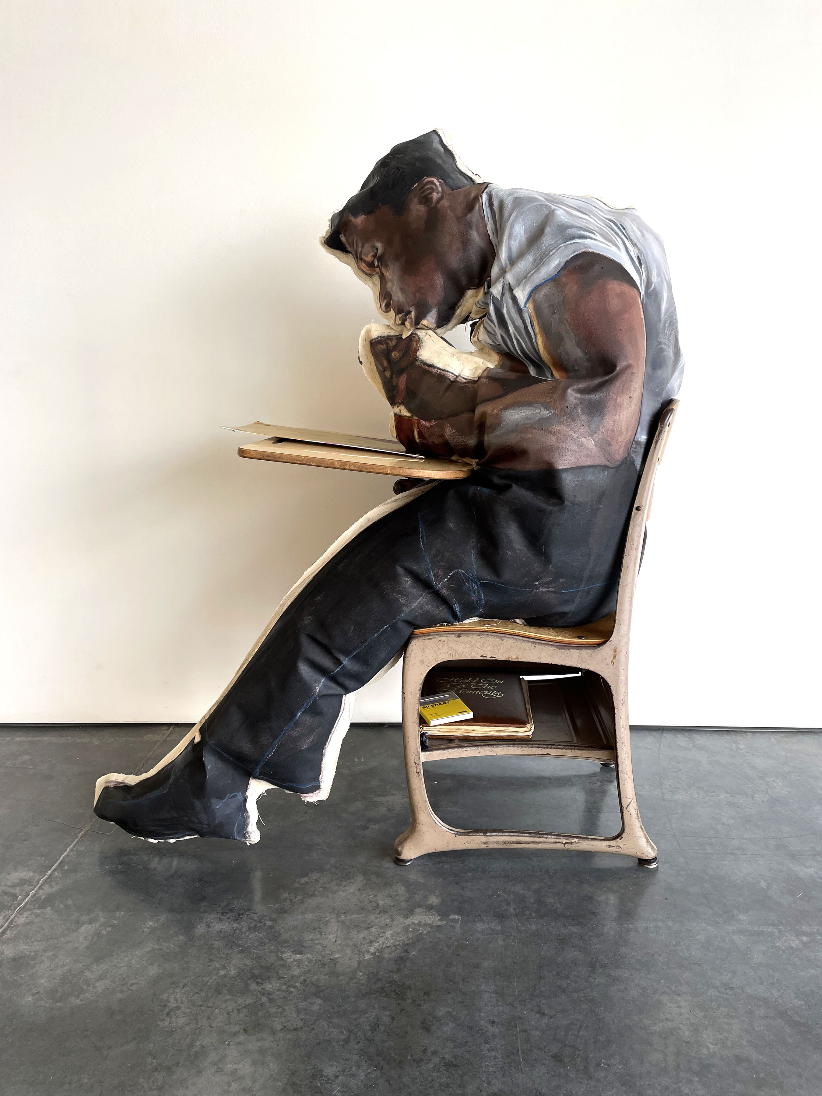 A hyperrealistic painted figure of a man seated on a wooden chair, hunched forward in a contemplative pose while reading from a wooden desk. The figure wears a light gray shirt and dark pants, rendered in oil paint with striking anatomical detail, set against a plain white wall and dark floor.