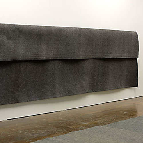 Large sheet of dark gray industrial felt suspended horizontally from a wall, with a pronounced fold creating a three-dimensional drape. The fabric hangs against a white wall above a wooden floor, emphasizing its weight and textural surface.