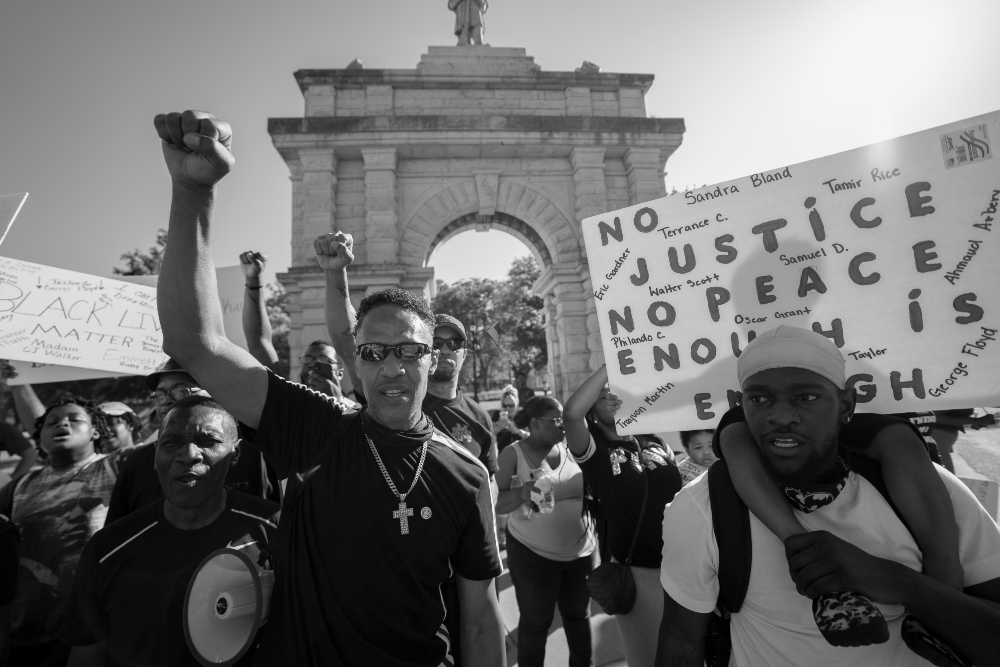 Black and white photograph of a protest gathering in front of a classical stone monument. In the foreground, a man wearing sunglasses and a cross necklace raises his fist while holding a megaphone, surrounded by other demonstrators. Visible protest signs include text reading "NO JUSTICE NO PEACE" and names of individuals. The composition captures the energy and solidarity of the crowd against the architectural backdrop.