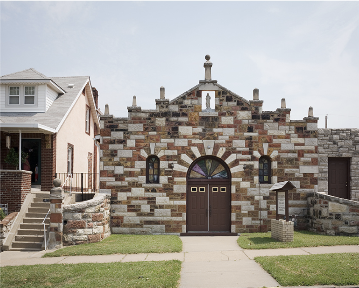 A stone church building with alternating red and cream-colored masonry dominates the composition, featuring a gabled roof topped with a cross and ornamental finials. Dark wooden double doors with a decorative fanlight occupy the center, flanked by arched windows, while a residential brick house with modern additions appears to the left of the frame.