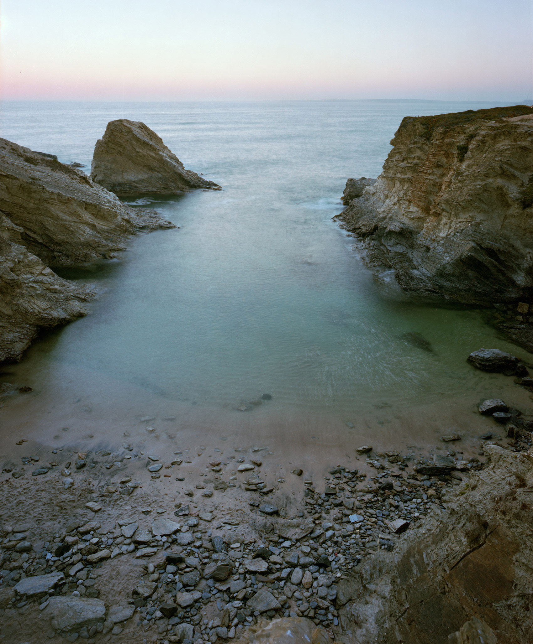 A rocky coastal cove at dusk, framed by steep limestone cliffs on either side. The smooth, milky water reflects the pale purple and blue tones of the twilight sky, while a solitary rock formation rises from the sea in the middle distance. The foreground shows a pebbly beach scattered with light-colored stones.