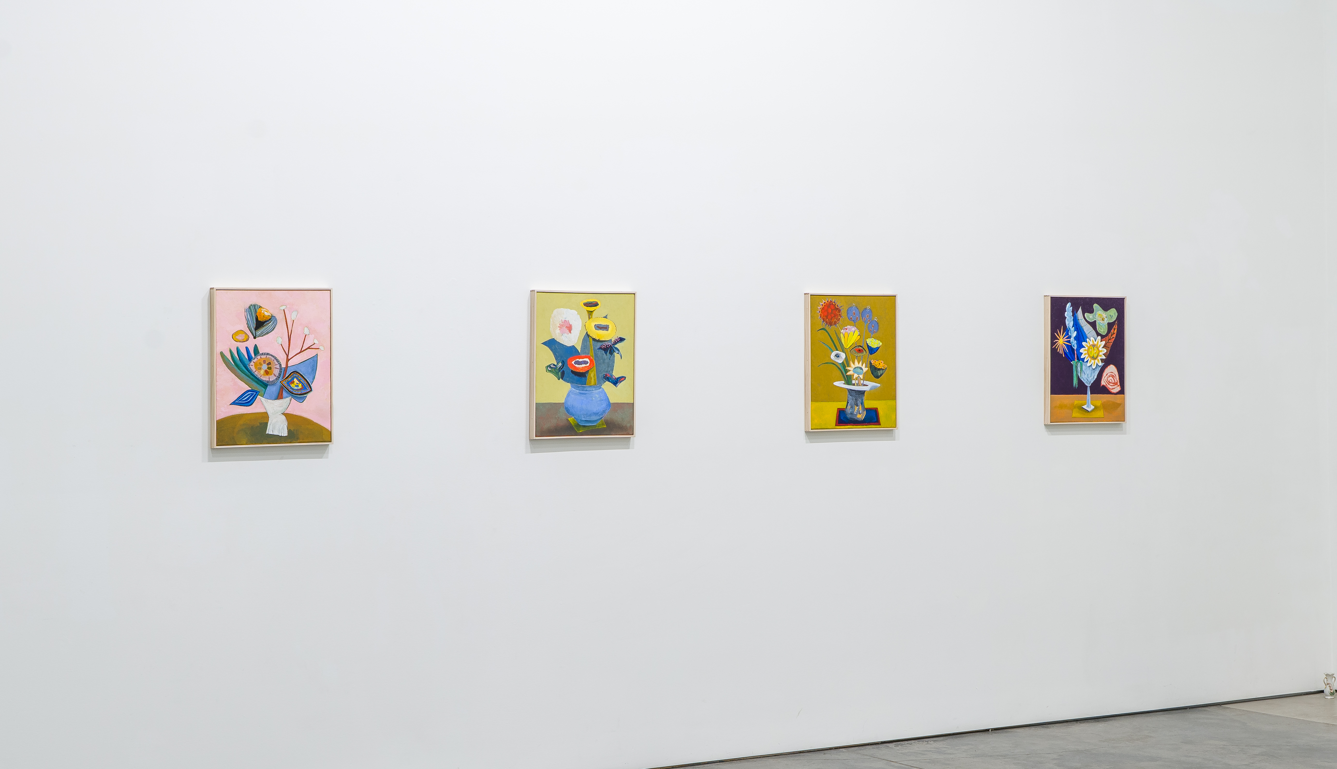 Four small framed paintings arranged in a horizontal line on a white gallery wall, each depicting stylized floral arrangements in ceramic vases against brightly colored backgrounds of pink, yellow, olive, and deep purple. The compositions feature bold, expressive brushwork with vibrant hues and imaginative botanical forms that blend recognizable and fantastical elements.
