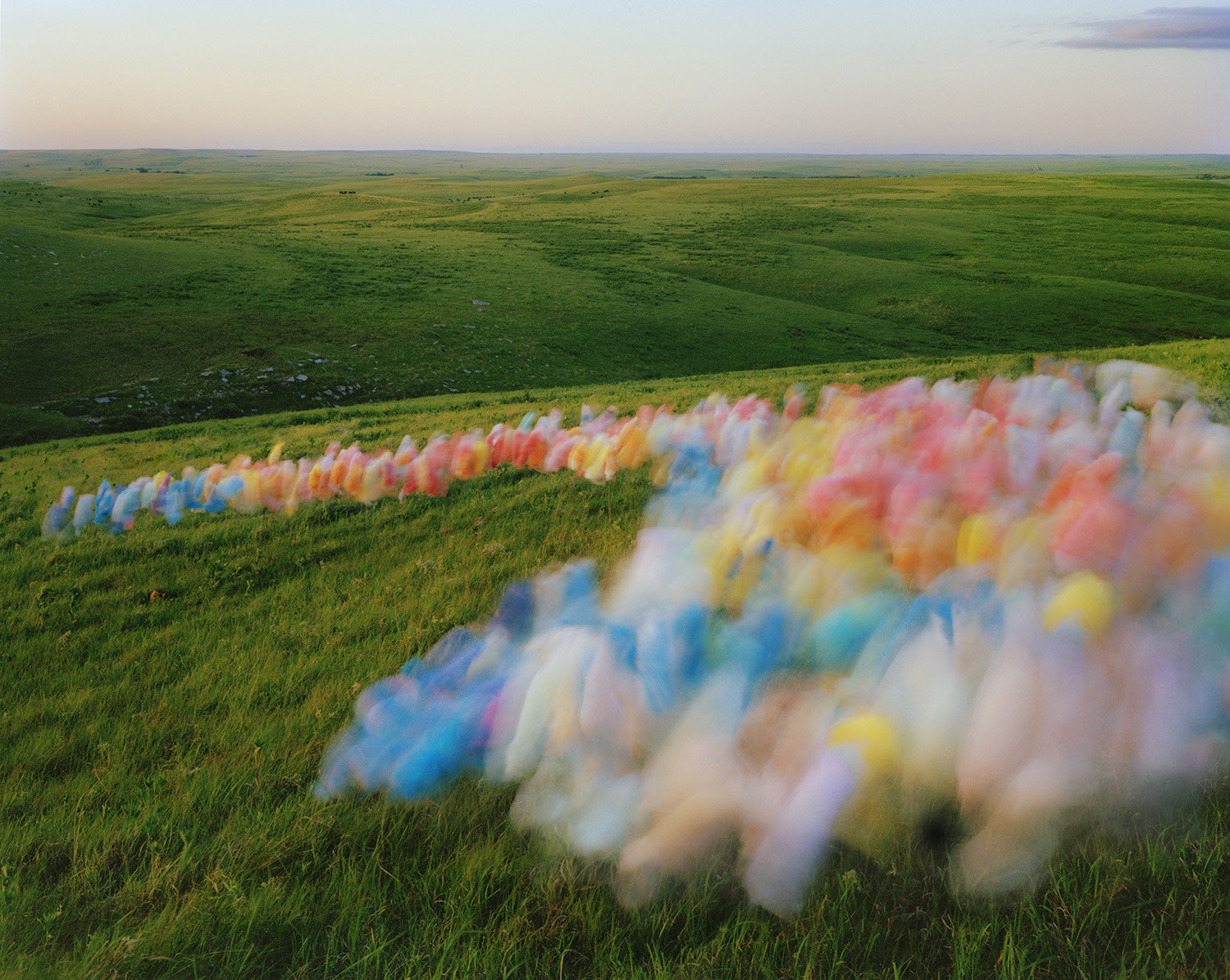 Blurred, multicolored tulle fabric arranged in undulating rows across rolling green prairie grassland under a pale sky, creating an abstract linear pattern that contrasts with the vast, natural landscape.