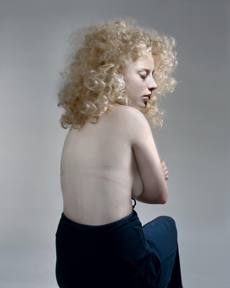 A pale-skinned figure with voluminous curled blonde hair is shown in profile against a neutral gray background, their torso bare and turned slightly toward the camera while their gaze is directed downward. They wear dark blue fabric draped low across their hips, creating a classical, sculptural composition with soft, luminous lighting.