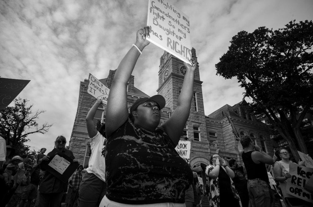 Black and white photograph of a protest scene featuring a central figure with raised fists holding a sign, surrounded by other demonstrators in front of a historic stone building with a clock tower. Additional protesters visible in the crowd hold various signs advocating for rights and justice.