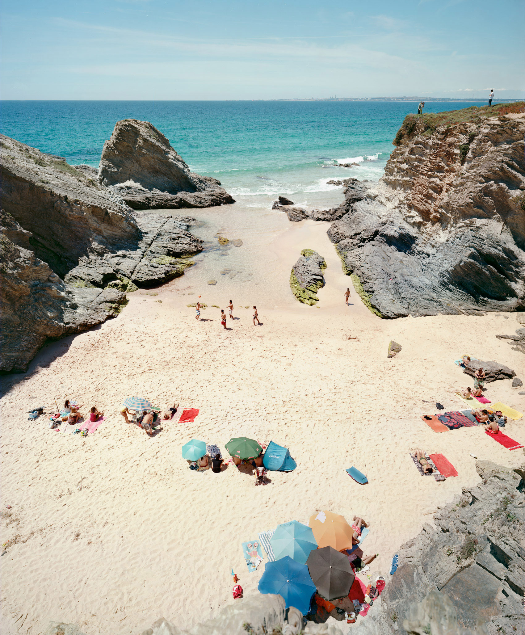 Aerial view of a secluded beach cove surrounded by dramatic rocky cliffs, with scattered beachgoers beneath colorful umbrellas and on towels dotting the pale sand. Turquoise water and white-capped waves frame the intimate sandy enclosure, while figures climb the cliffsides and populate the beach in small groups.