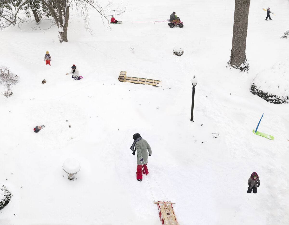 Aerial view of a snowy hillside dotted with people engaged in winter activities—sledding, playing, and walking—scattered across the white landscape with bare trees, a lamp post, and various sleds and toys visible from above.