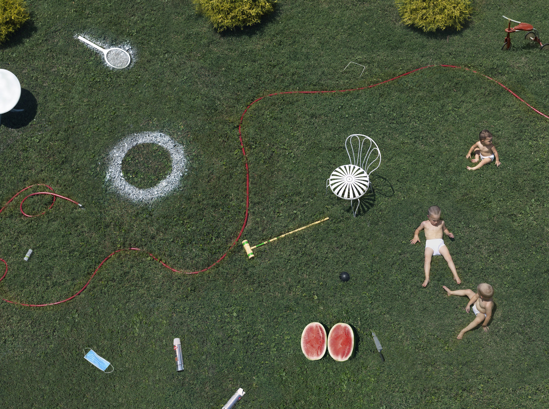 Aerial view of a lush green lawn scattered with everyday objects and three shirtless children at play. White chalk circles, a red garden hose, lawn furniture, a watermelon cut in half, gardening tools, and household items are arranged across the grass in a carefully composed domestic scene.