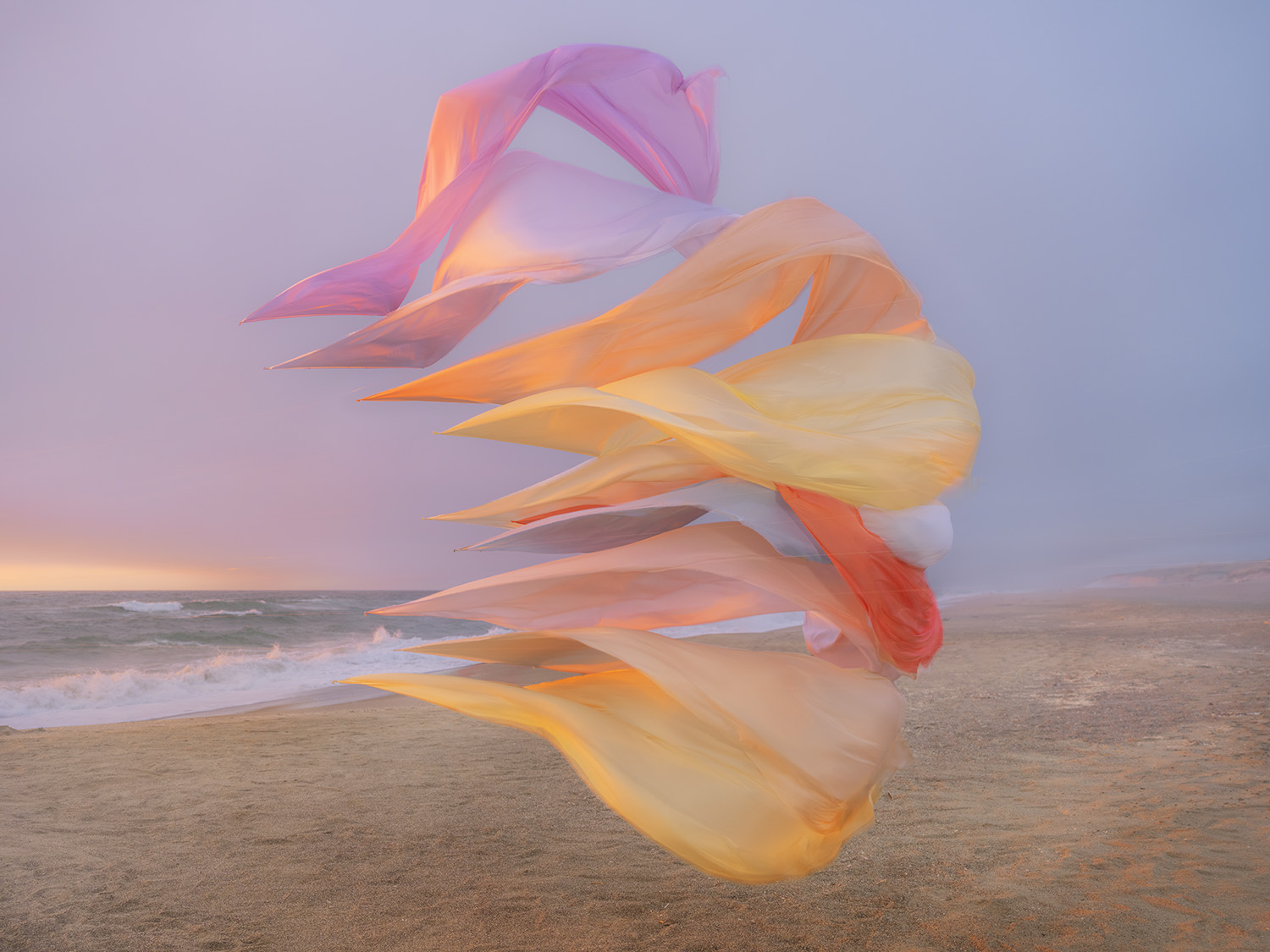 Billowing silk ribbons in shades of pink, orange, and yellow flow dynamically across a sandy beach at sunset, with ocean waves and a dusky sky visible in the soft-focus background.