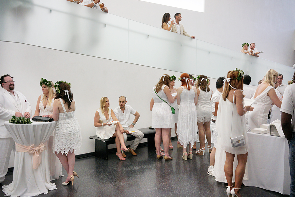 A large group of people dressed in white clothing gather in a modernist interior space with white walls and a dark floor. Guests mingle and socialize across multiple levels, with some standing on an elevated platform visible in the background, while others interact in the main hall below. Green floral garlands and white-draped cocktail tables accent the scene.