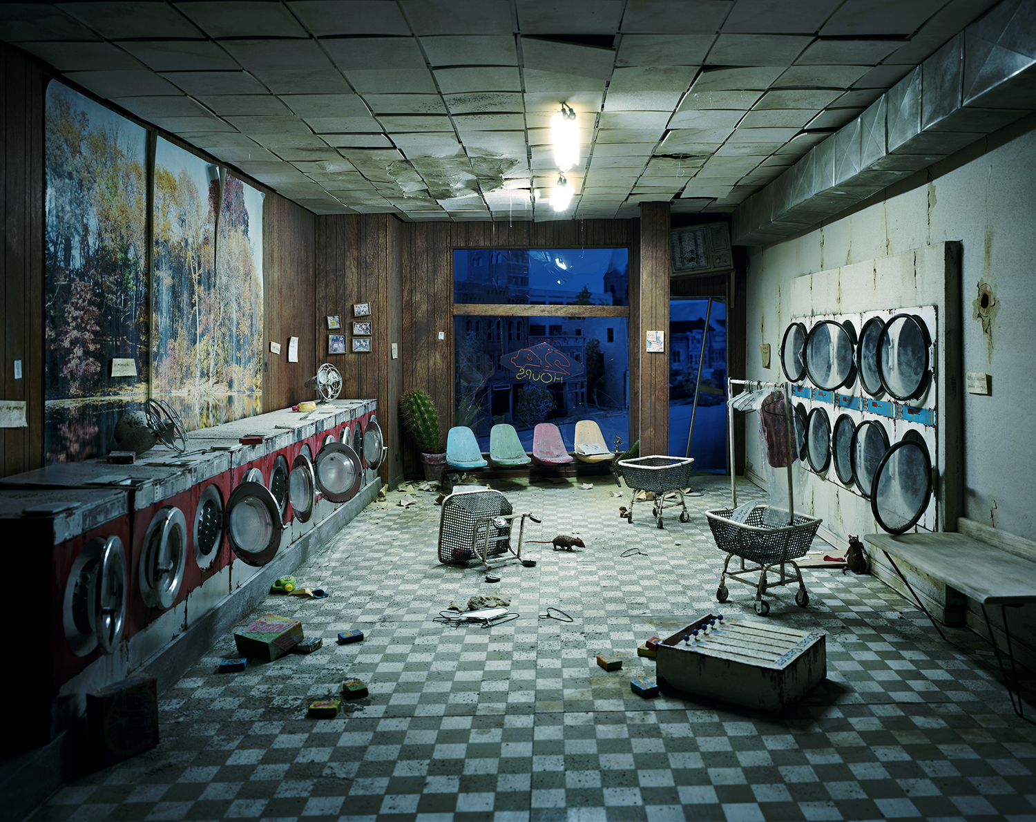 An abandoned laundromat interior at dusk, with rows of red and white washing machines lining the left wall and dryers mounted on the right. Four pastel-colored chairs sit near the center, while laundry baskets, scattered clothing, and debris cover the checkered floor. Large weathered murals decorate the back wall beside a blue-tinted storefront window, creating an eerie, desolate atmosphere beneath flickering fluorescent lights.