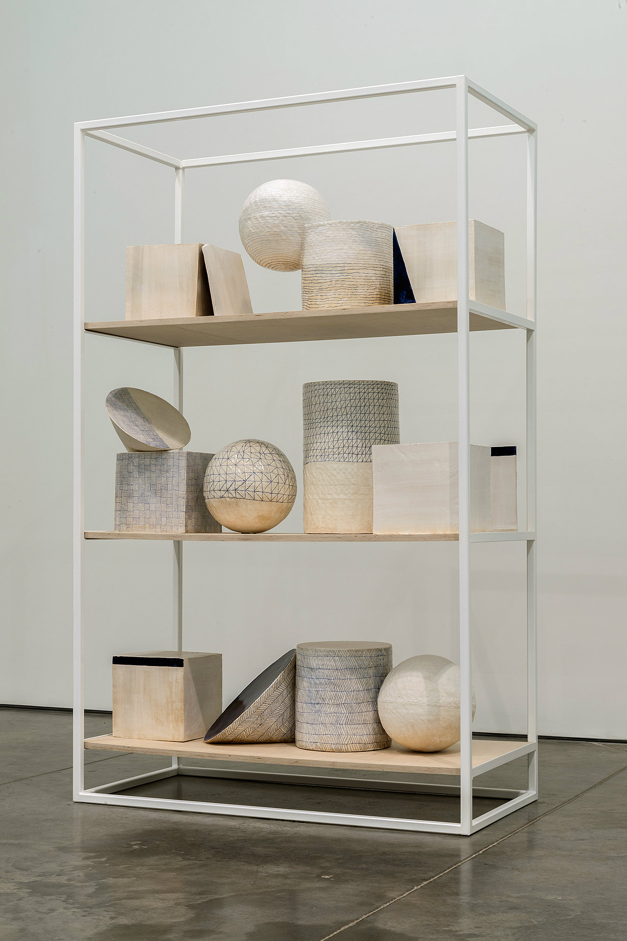 White steel display case with four wooden shelves containing an arrangement of geometric sculptural objects in natural materials including woven baskets, spheres, cylinders, and rectangular forms in cream, beige, gray, and black tones.