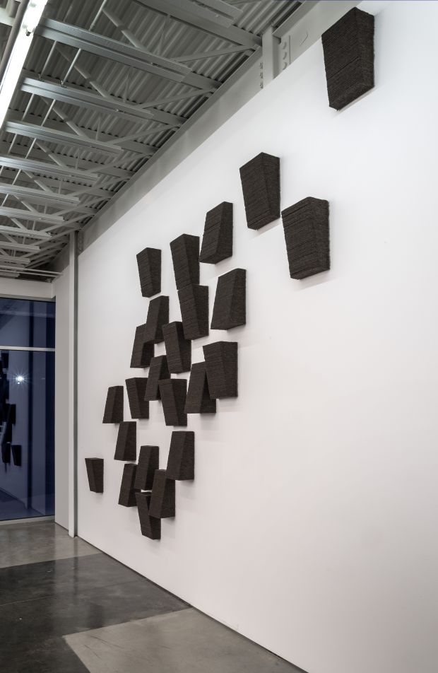 A composition of approximately twenty-four dark charcoal-colored rectangular forms mounted on a white gallery wall in an ascending diagonal arrangement. The textured, three-dimensional objects cast subtle shadows and vary slightly in size and orientation, creating a dynamic kinetic effect across the expansive white surface.