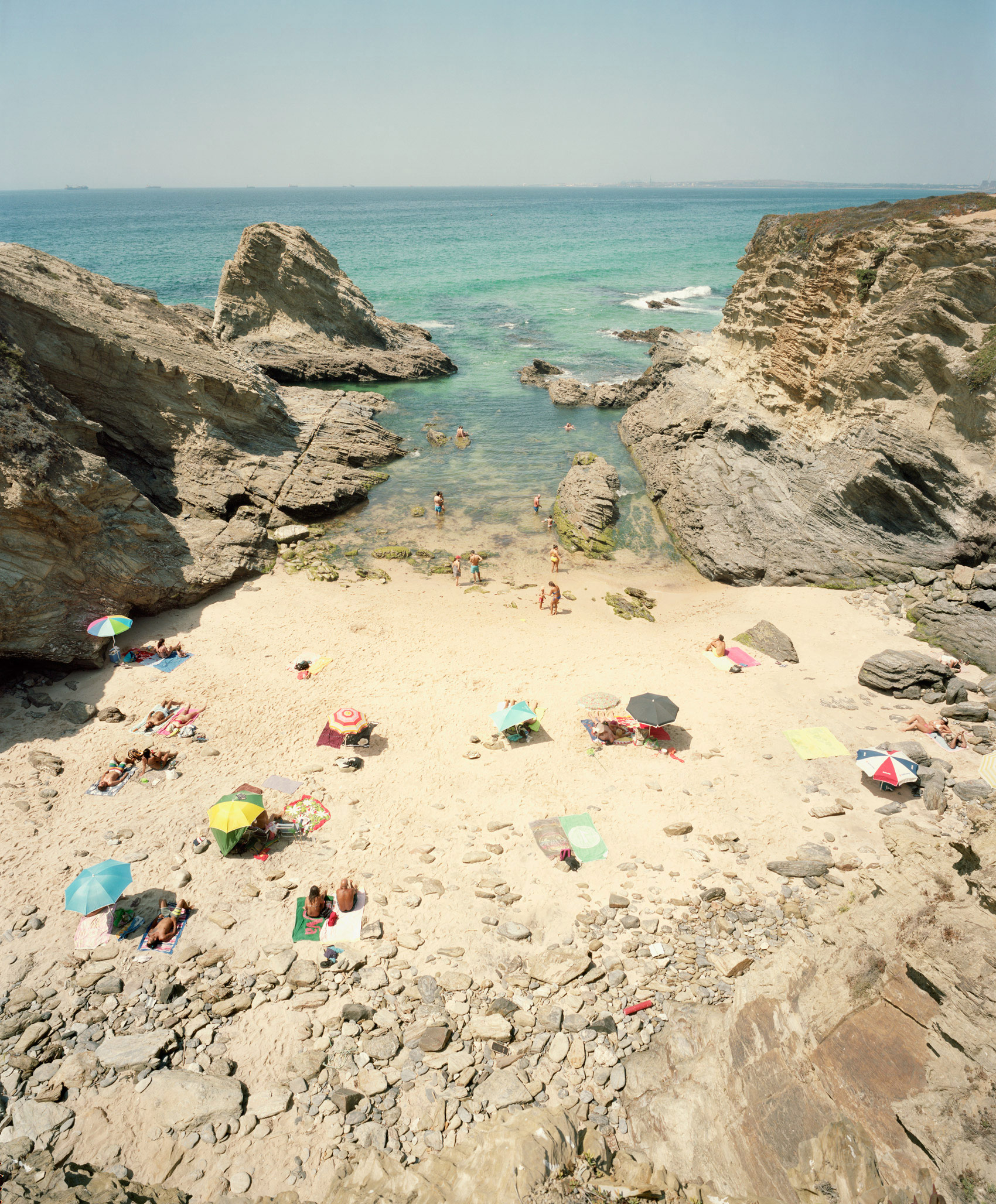 Aerial view of a secluded cove beach framed by dramatic limestone cliffs, with colorful umbrellas and sunbathers scattered across pale sand, turquoise water visible between rocky outcrops, and a clear horizon stretching across the composition.