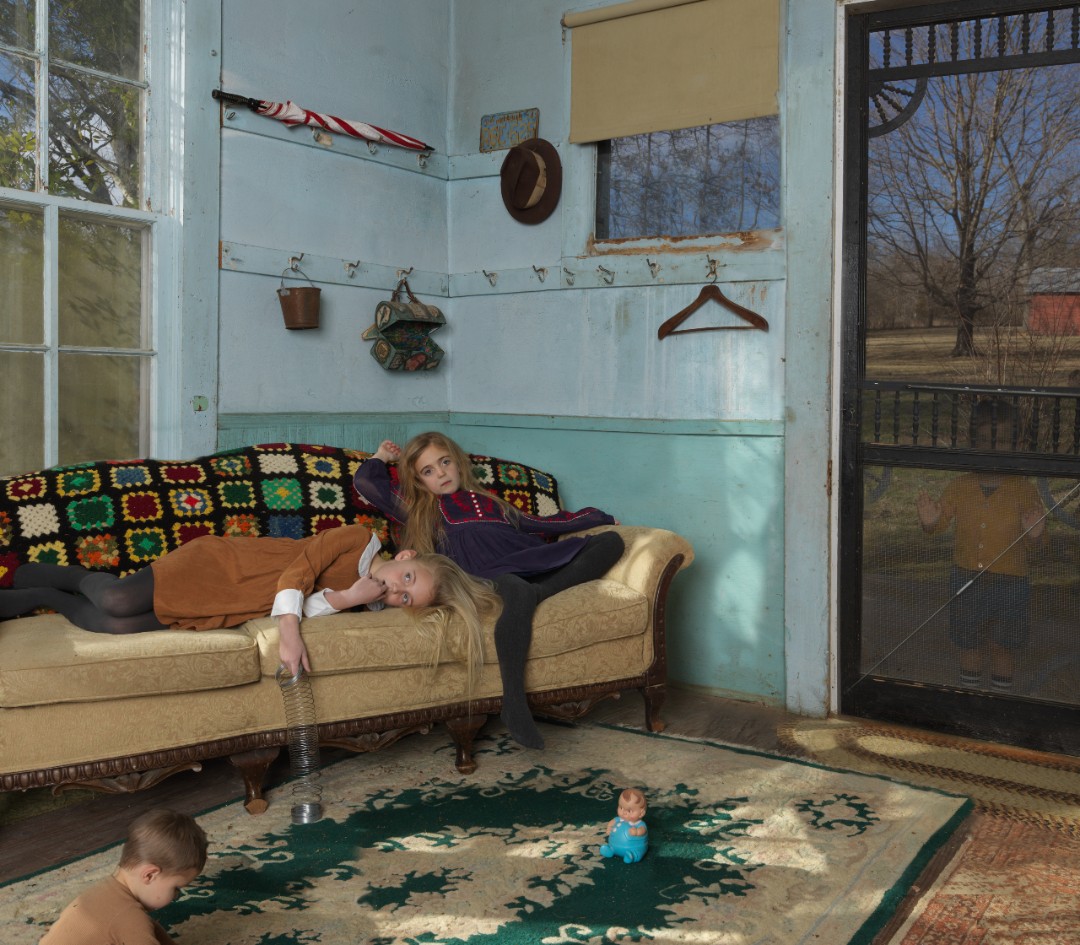 An interior scene of a light blue-walled room with a woman and two children occupying a worn upholstered sofa and floor. A young girl lies across the cushions wrapped in a colorful knit blanket while a woman reclines behind her; a boy sits on the floor beside a patterned rug, and a small child in blue sits in the center of the rug. Sunlight streams through windows revealing bare trees outside, illuminating hooks and domestic objects hung on the walls and casting warm shadows across the space.