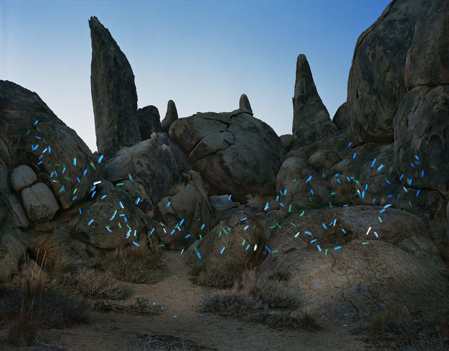 Desert rock formation with tall granite boulders and pinnacles against a clear blue sky, overlaid with bright blue and green painted or digital file tab shapes scattered across the sandy ground and lower rock surfaces.