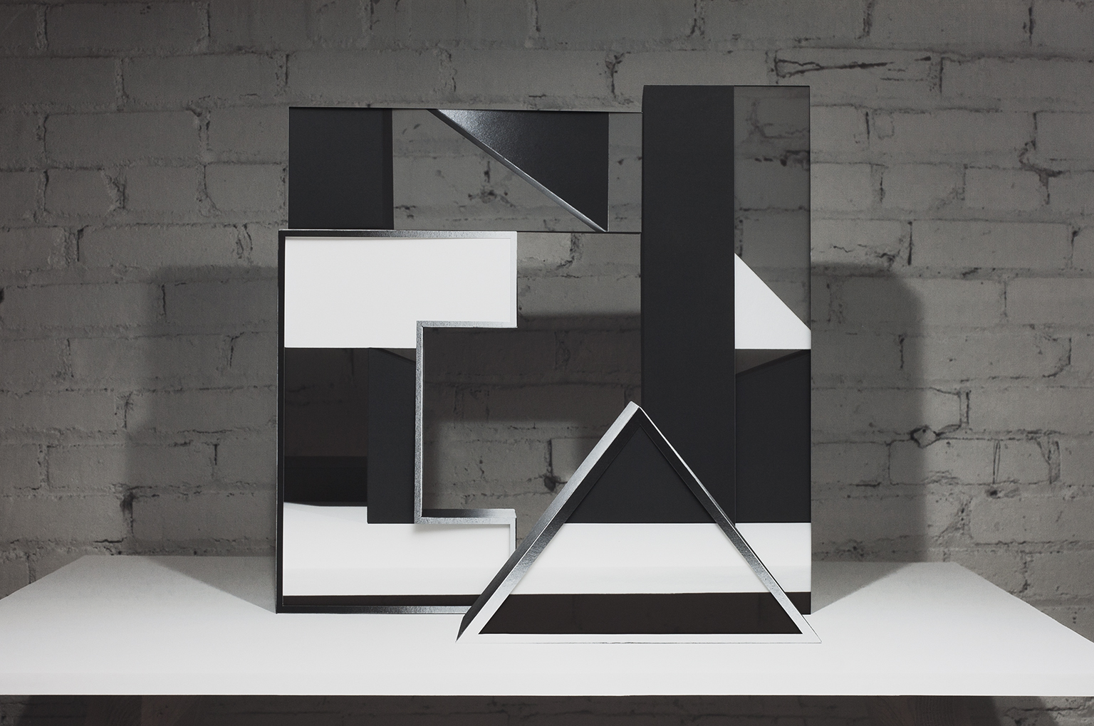 Abstract geometric composition of layered black and white paper forms arranged on a white platform against an exposed brick wall. Interlocking rectangles and a triangular shape create a three-dimensional collage with sharp angles and negative space, emphasizing contrast between dark and light surfaces.