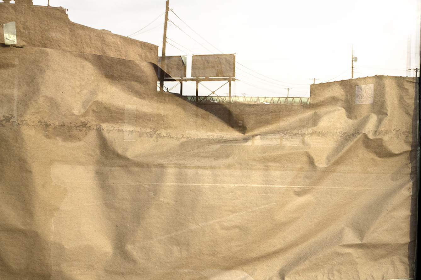 A layered photograph depicting an industrial sand or aggregate storage facility with sloped mounds of tan-colored material. Brick buildings with water tanks sit atop the landscape, connected by metal structures and utility poles. The composition creates a flattened, almost abstract quality through overlapping translucent photographic planes.