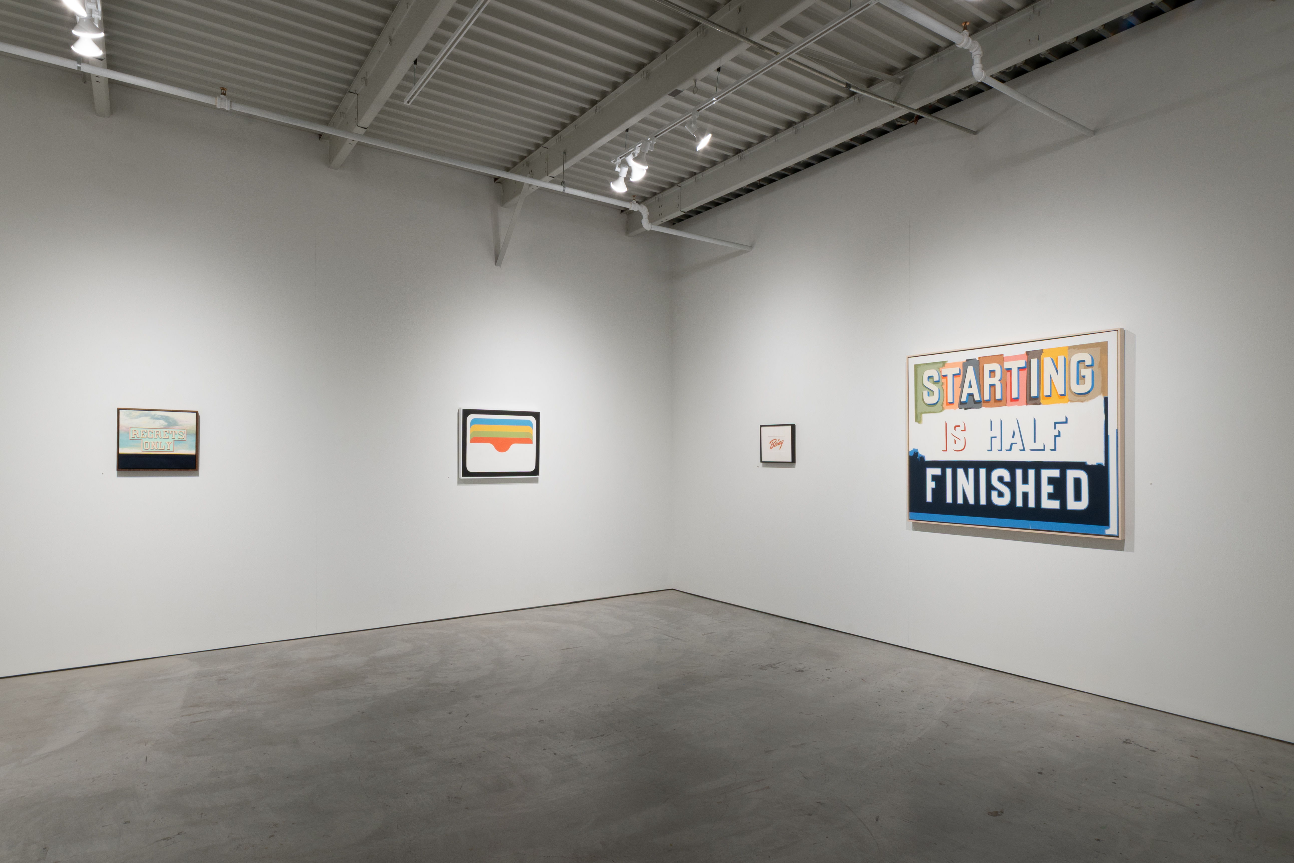 A gallery wall displaying four framed artworks of varying sizes against white walls and polished concrete flooring, with industrial ceiling infrastructure visible above. The largest work on the right features bold text reading "STARTING IS HALF FINISHED" in yellow, white, and blue lettering on a dark background. The remaining three smaller framed pieces are arranged across the left and center walls, including what appears to be landscape imagery and colorful geometric forms, each with simple black frames.