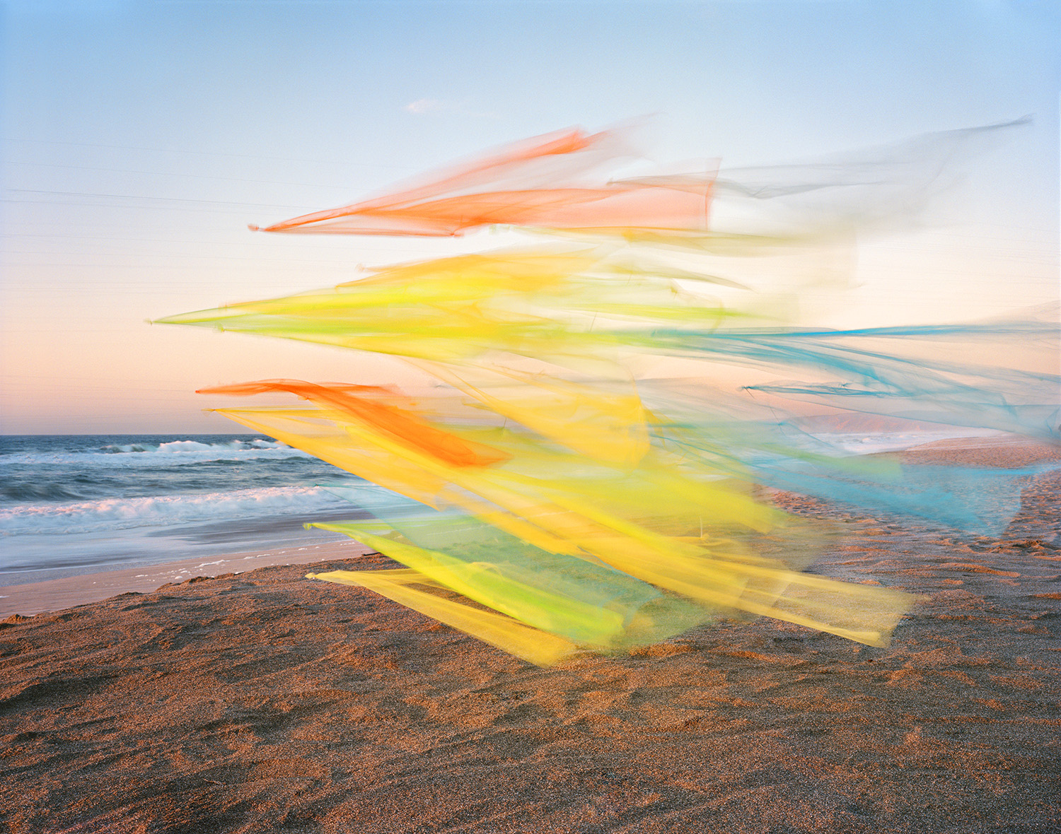 Translucent, flowing ribbons of yellow, orange, and blue tulle billow across a sandy beach at sunset, with gentle waves and a twilight sky visible in the background.
