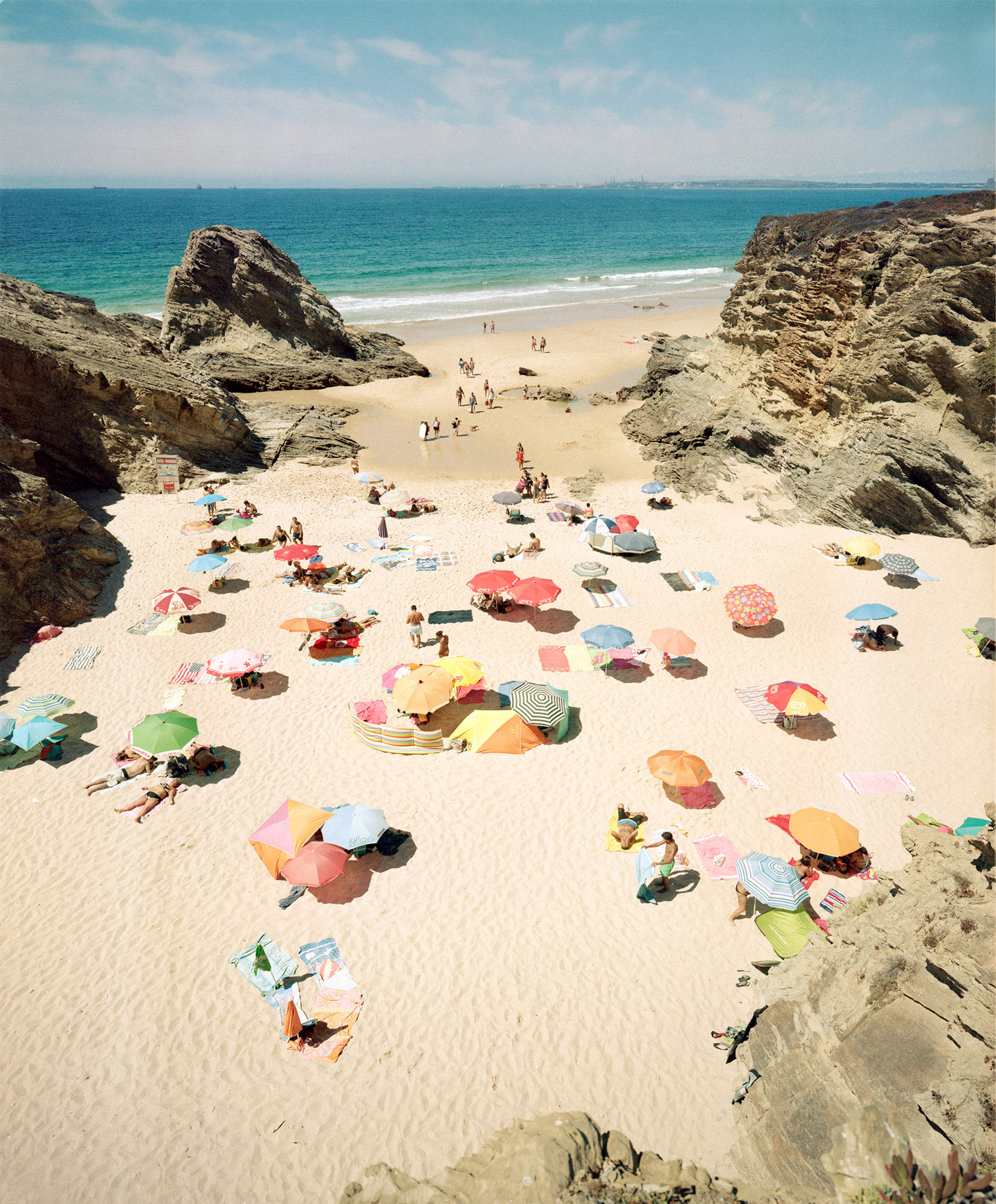 Aerial view of a crowded beach nestled between dramatic rocky cliffs, with beachgoers scattered across pale sand beneath a vibrant array of colorful umbrellas in red, yellow, blue, pink, and striped patterns. Turquoise waters and white-sand shoreline extend to the horizon under a partly cloudy sky.