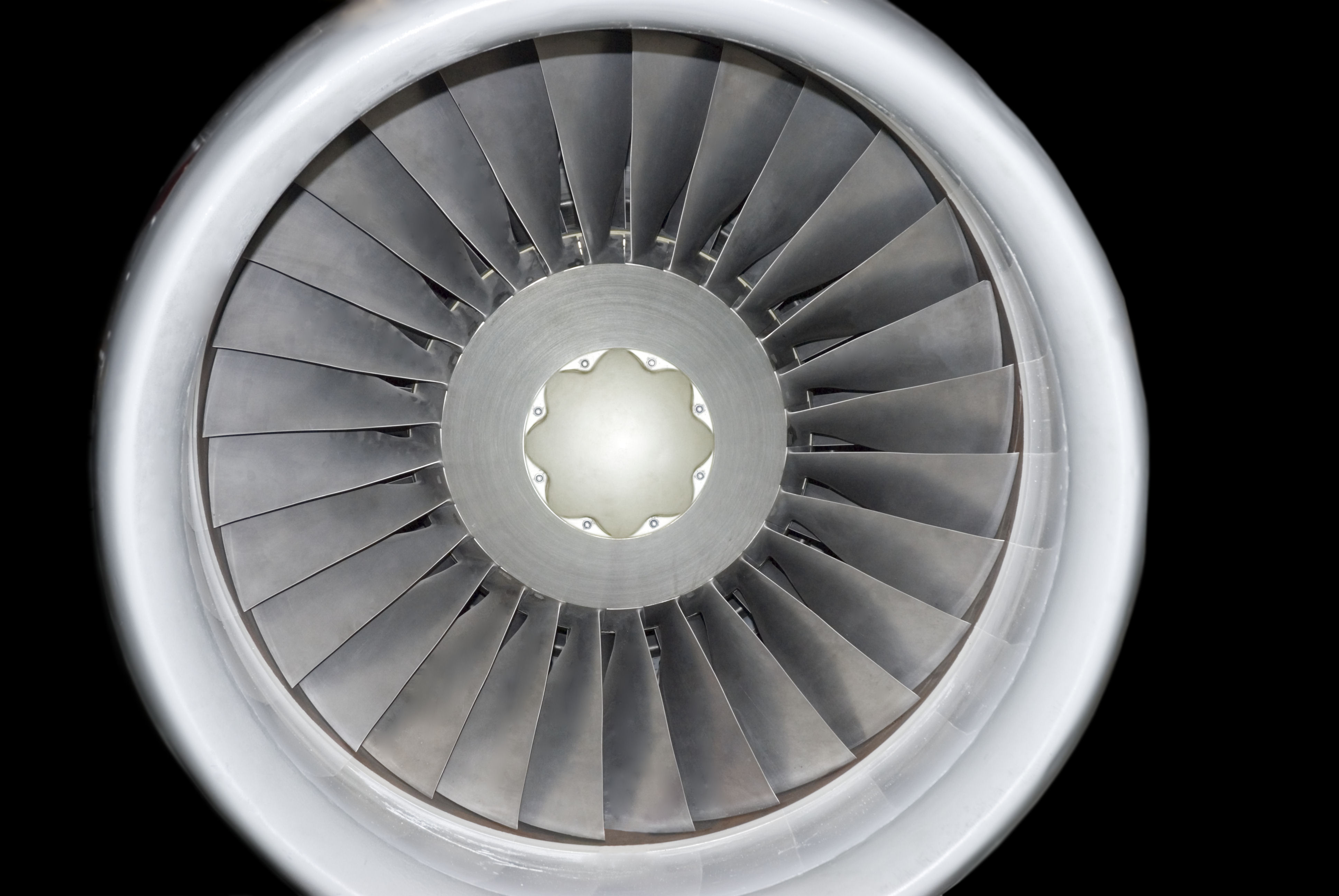 Front-facing view of a jet engine turbine with concentric metal blades radiating from a central hub, rendered in grayscale tones against a black background. The precise, symmetrical composition emphasizes the industrial geometry and reflective surfaces of the machinery.