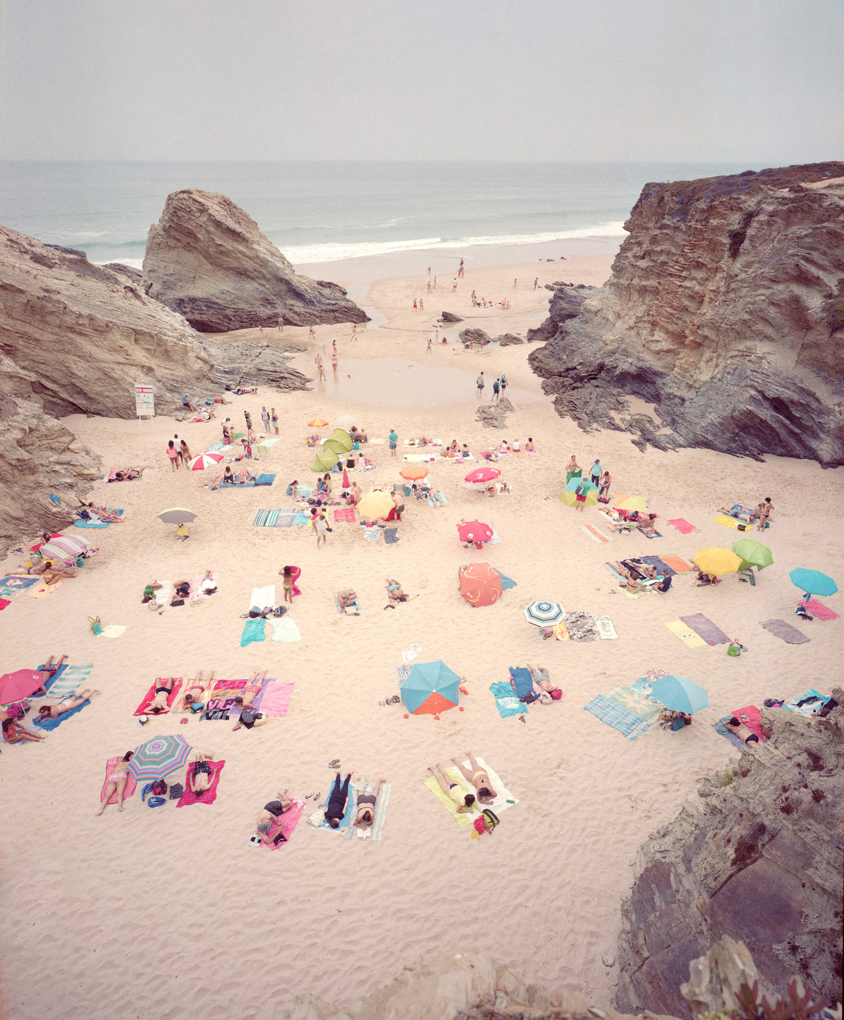 Aerial view of a crowded beach cove nestled between dramatic rocky cliffs, with colorful umbrellas, towels, and beachgoers scattered across pale sand, creating a vibrant patchwork of reds, pinks, yellows, blues, and greens against the neutral landscape.
