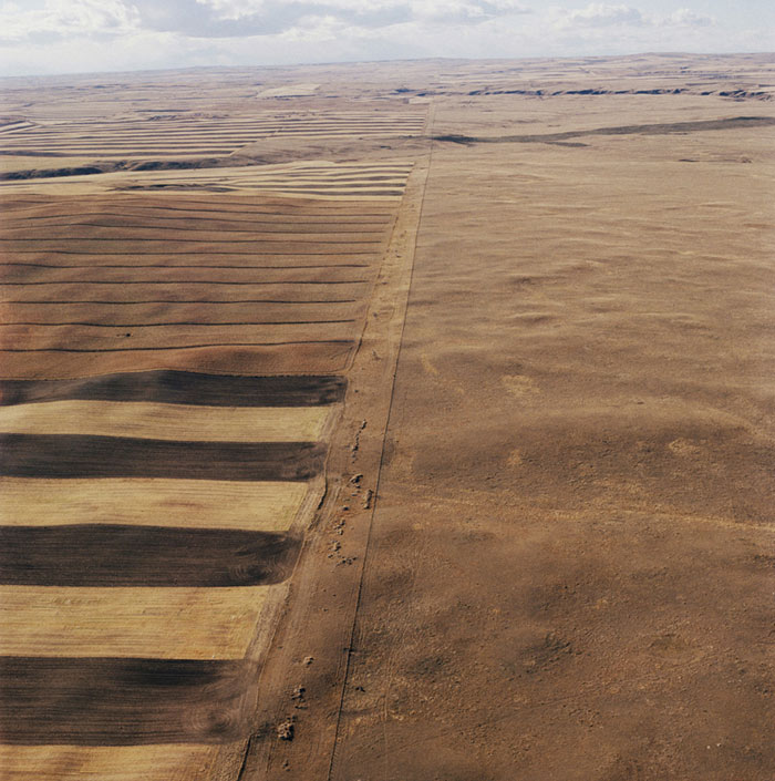 Aerial view of agricultural land divided by a straight dirt road, with plowed fields in varying shades of brown and tan on the left and an open, fallow field on the right. The flat, expansive landscape stretches toward the horizon beneath a cloudy sky.